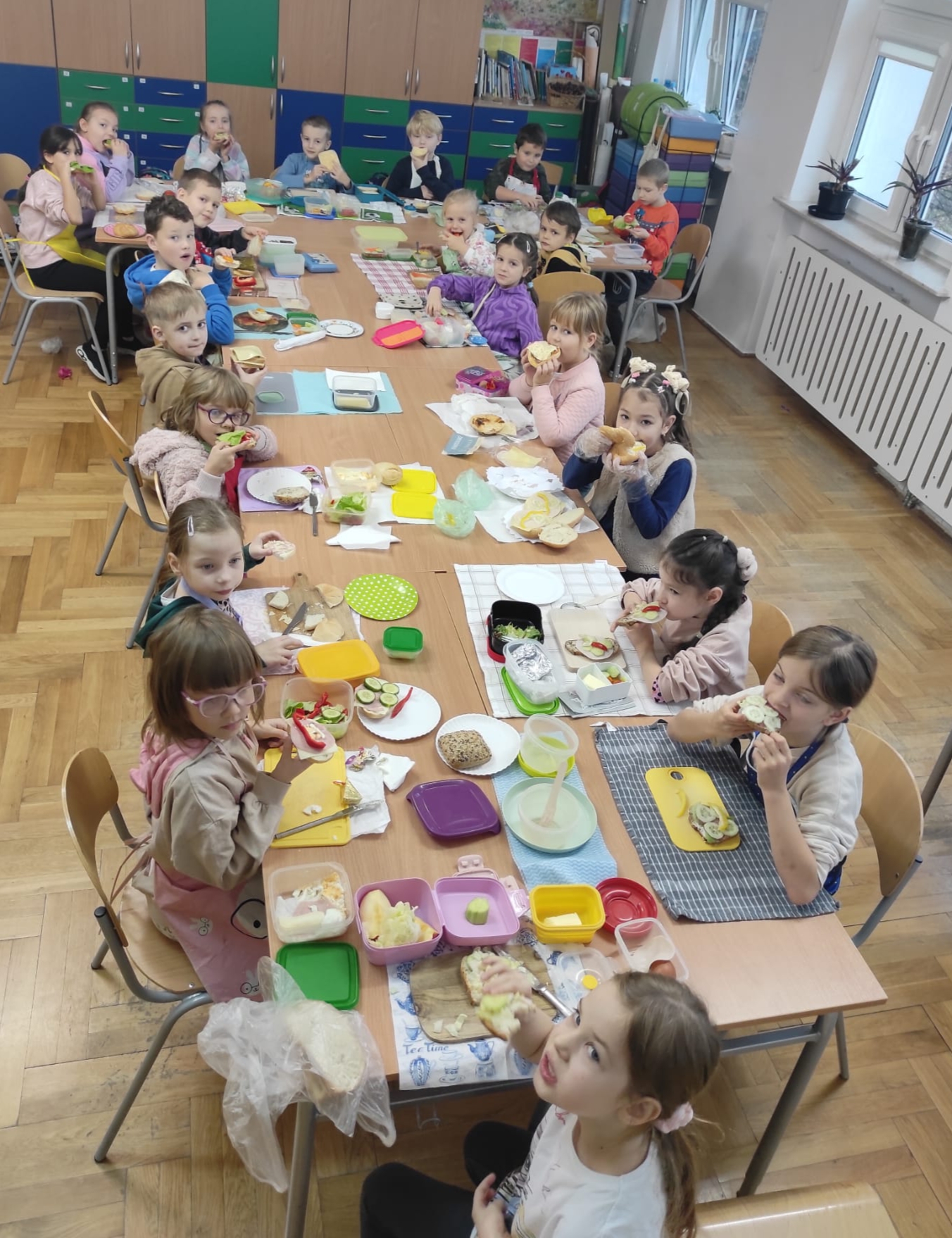 21 dzieci siedzi w sali lekcyjnej dookoła dużego stołu i samodzielnie przygotowują sobie kanapki. Przed nimi leżą pojemniki z różnymi produktami tj. chleb, masło, wędliny i warzywa oraz tacki i serwetki.