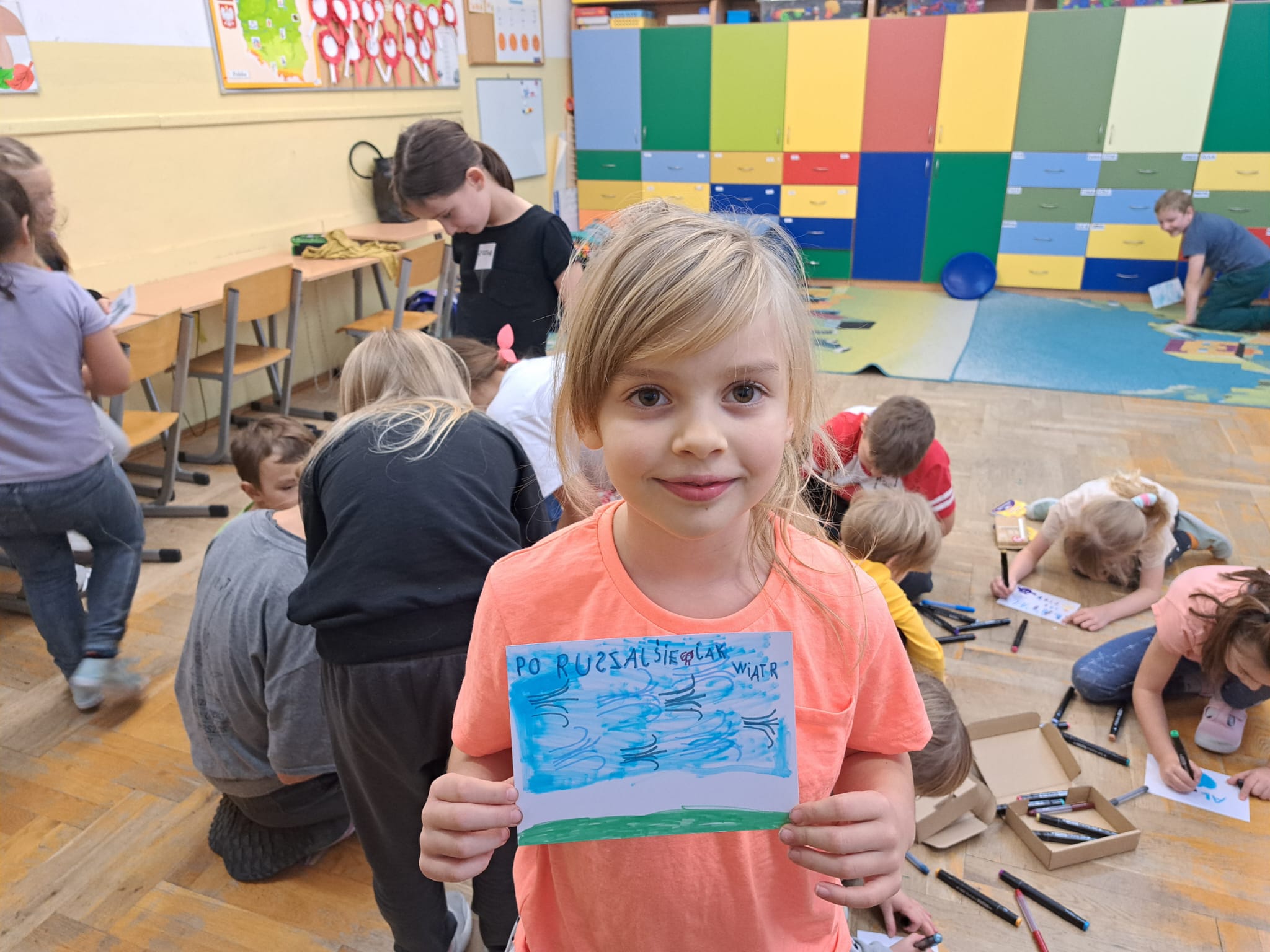 Na pierwszym planie dziewczynka uśmiecha się i trzyma w rękach rysunek z napisem „PO RUSZASZ SIĘ JAK WIATR”. W tle inne dzieci rysują i kolorują, siedząc na podłodze. W klasie znajdują się kolorowe szafki oraz dekoracje edukacyjne na ścianach. 