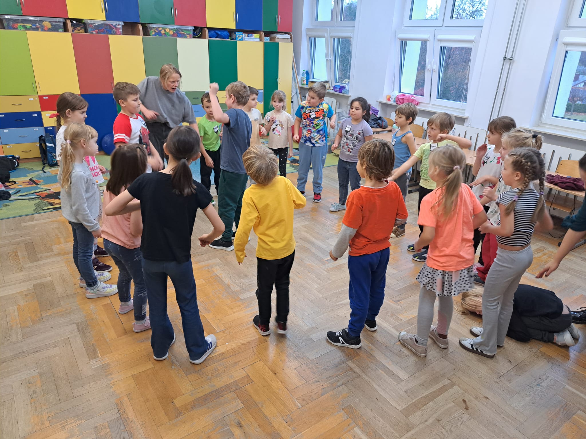 Grupa uczniów wykonuje ćwiczenia ruchowe, stojąc w kręgu. Naśladują chłopca, który stoi w środku kręgu. W tle widać kolorowe szafki, a na ścianach znajdują się mapy i dekoracje edukacyjne. Sala jest jasna i przestronna, a atmosfera wygląda na radosną i energiczną.