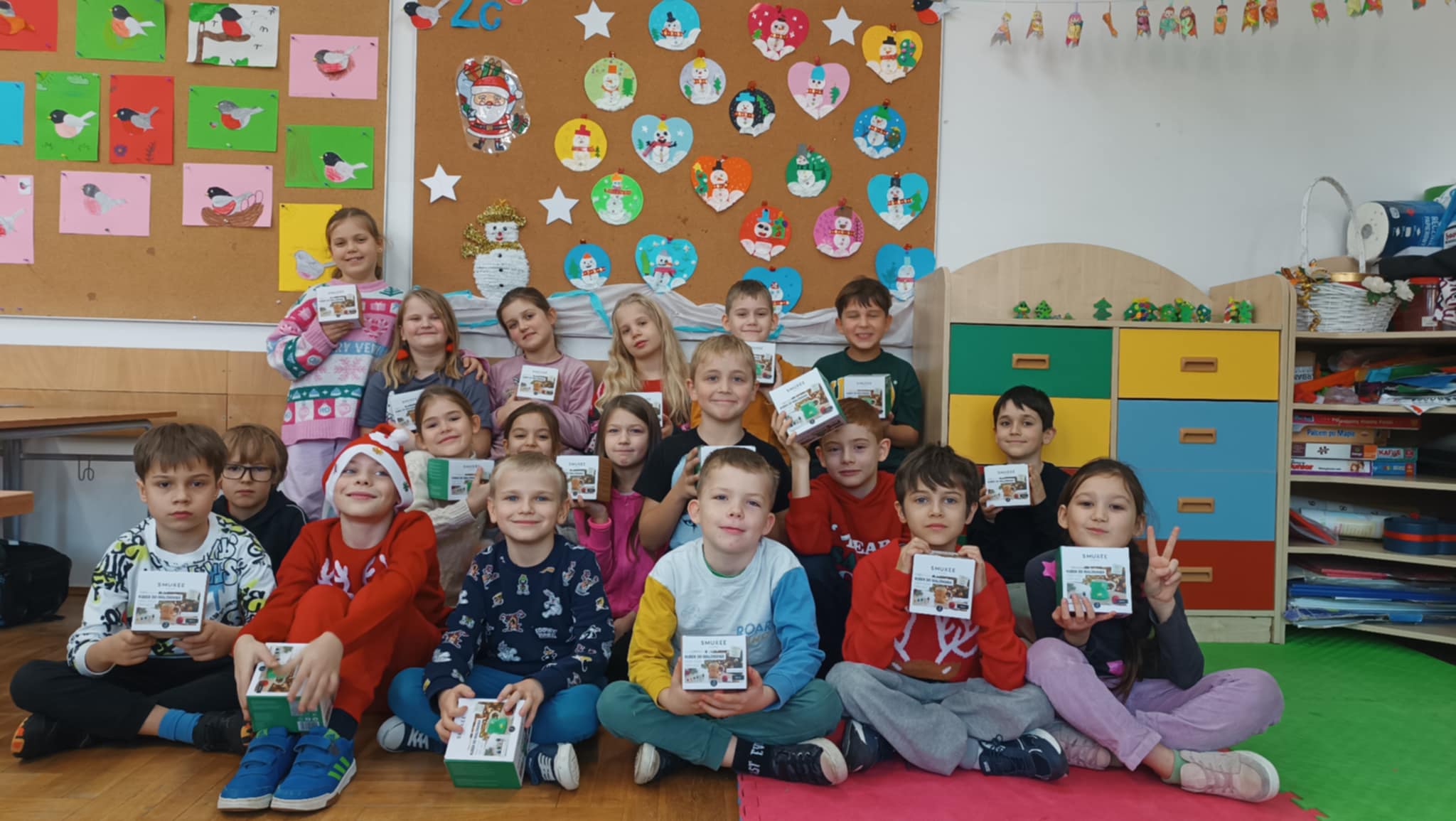 Klasa szkolna w świątecznym nastroju. Dzieci trzymają prezenty, prawdopodobnie otrzymane w ramach świątecznej akcji lub uroczystości. W tle widoczne są świąteczne ozdoby i dziecięce prace plastyczne.