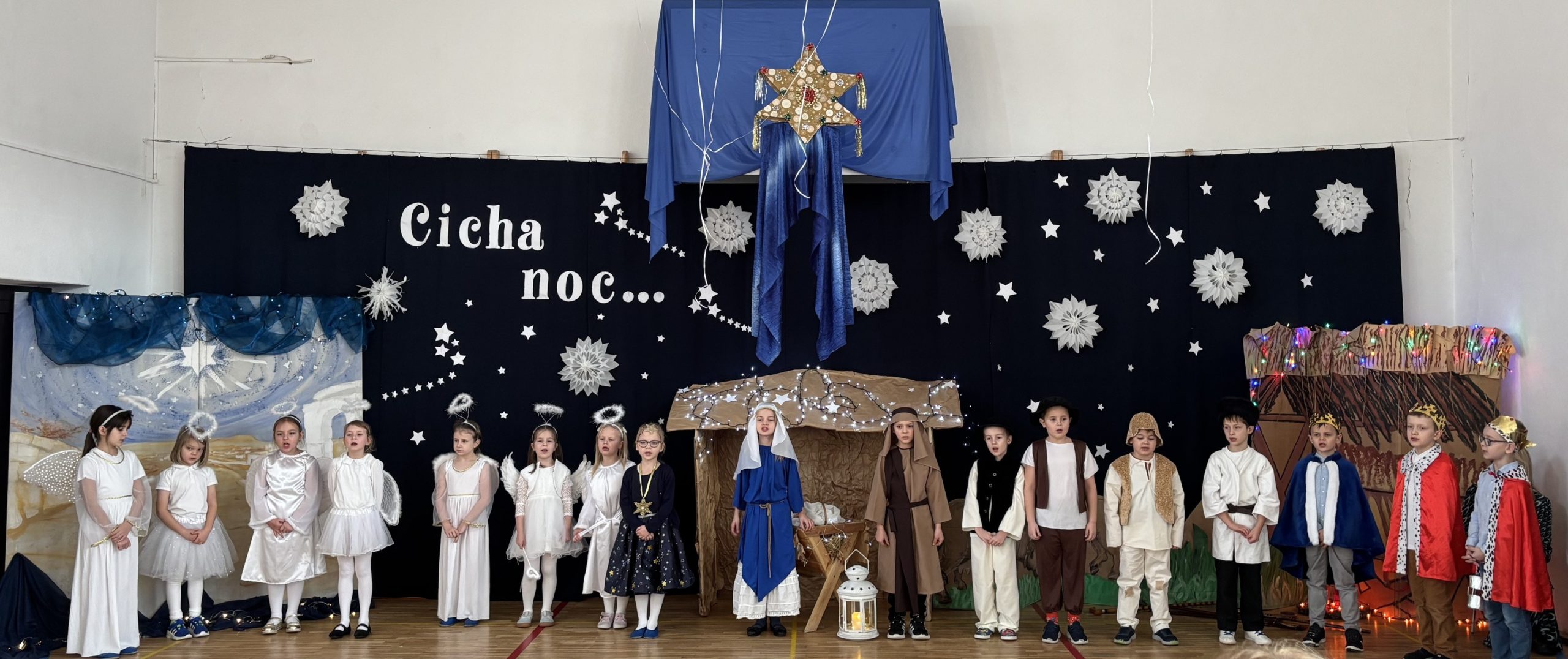 W sali gimnastycznej na tle dekoracji świątecznej z napisem „Cicha noc...”, stoi grupa uczniów ubrana w stroje pastuszka, aniołka, królowie.