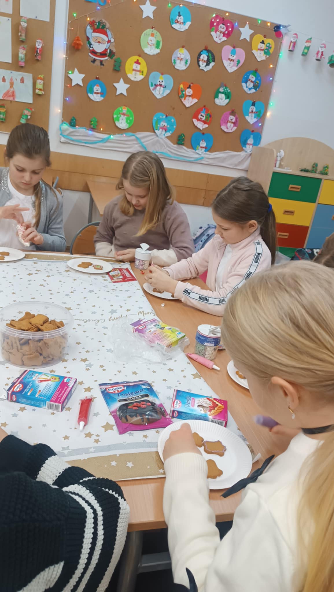 Grupa dziewczynek siedzi przy stole i dekoruje pierniczki lukrem i posypkami. Na stole znajdują się pojemniki z pierniczkami, lukrem i ozdobami. W tle widoczna jest tablica ze świątecznymi dekoracjami.