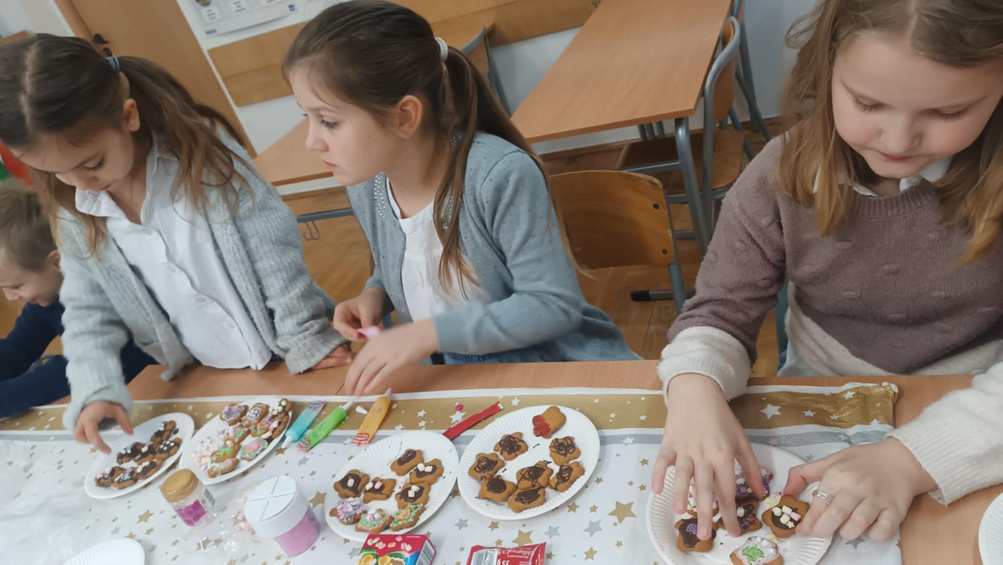 Dzieci skupione na dekorowaniu świątecznych pierniczków. Używają lukru, kolorowych posypek i innych ozdób tworząc unikalne wzory.