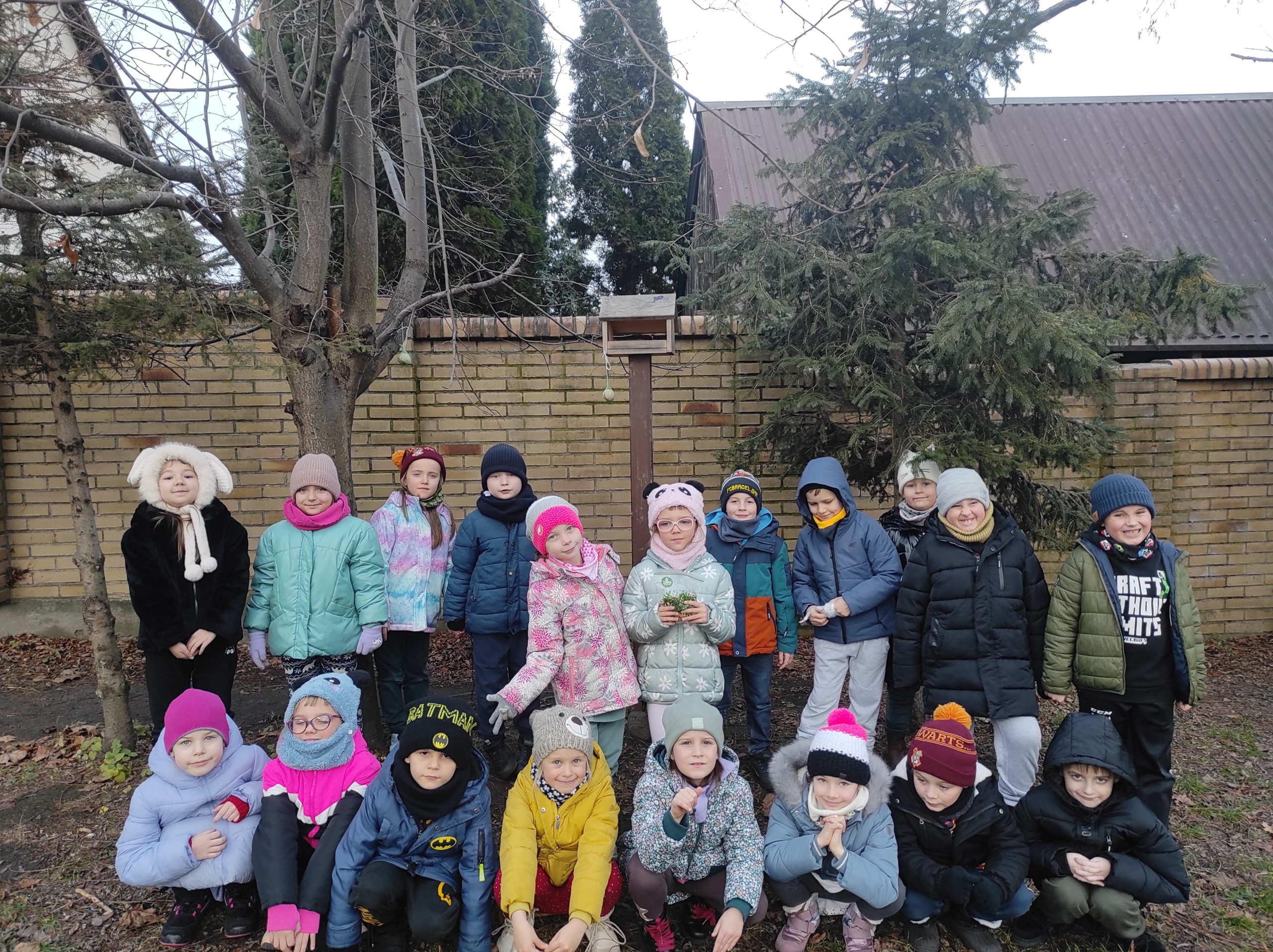 Uczniowie stoją w dwóch rzędach, jeden stoi, drugi kuca. Jedna stojąca dziewczynka trzyma coś w dłoniach.  W tle fragment muru, dwa drzewa. Pomiędzy drzewami widoczna jest kantówka, do której przytwierdzony jest karmnik. Na drzewie i przy karmniku wiszą dwie kule tłuszczowe dla ptaków.