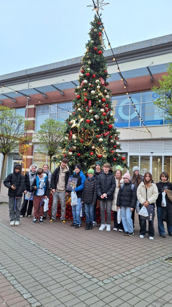 Grupa uczniów stoi przy wielkiej choince ozdobionej świątecznymi ozdobami. Wszyscy ubrani w ciepłe kurtki trzymają w rękach upominki. Są uśmiechnięci