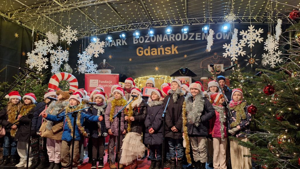 Duża grupa dzieci stoi na scenie. Na głowie mają czapki św. Mikołaja, a na szyjach zawieszone łańcuchy choinkowe. Wokół nich są ozdobione choinki, a z góry zwisają sztuczne duże płatki śniegu. Z tyłu jest napis Jarmark Bożonarodzeniowy Gdańsk.