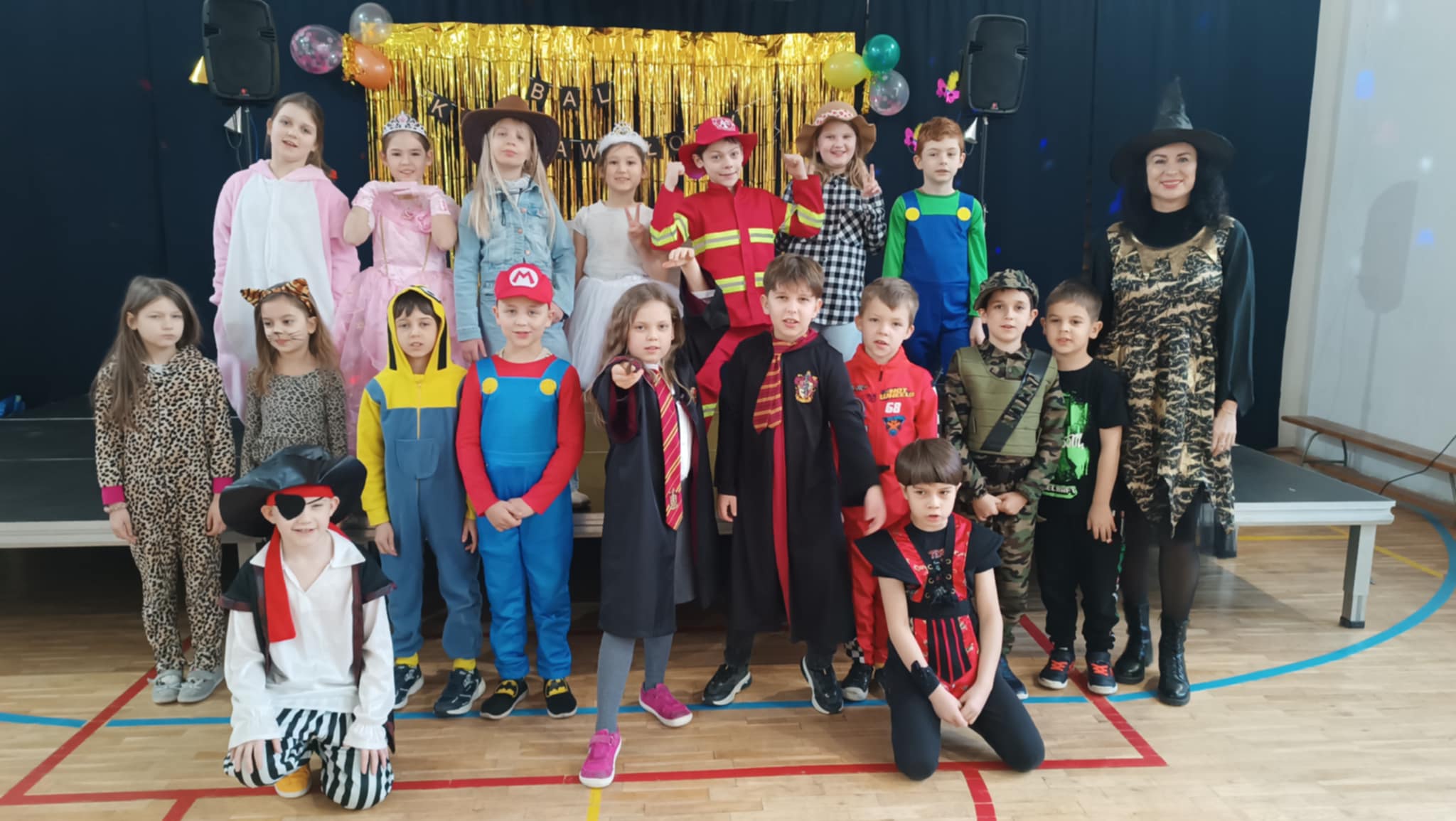 Grupa dzieci w wieku szkolnym przebranych w różnorodne stroje kostiumy, takie jak super bohaterowie, postacie z bajek stoi w dwóch rzędach pozując do zdjęcia na scenie. Nauczycielka w stroju czarownicy stoi po prawej stronie z tyłu.