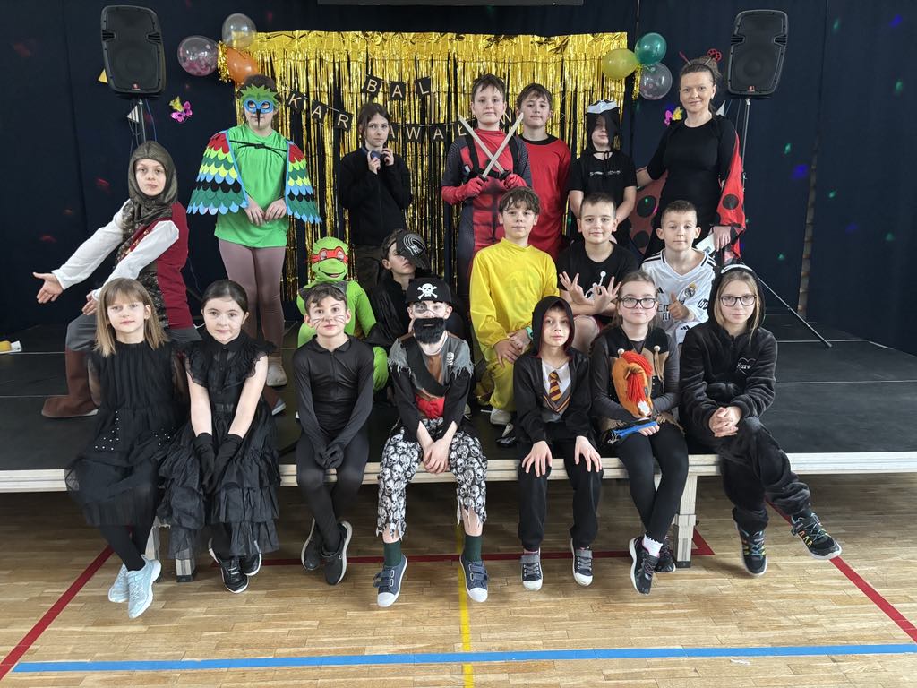 W sali gimnastycznej na podeście, na tle ciemnego płótna i karnawałowej dekoracji, pozuje do zdjęcia osiemnastu przebranych w karnawałowe stroje uczniów klasy 3c wraz z wychowawczynią.