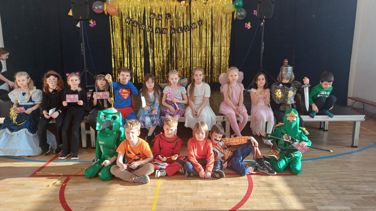 Grupa 18 dzieci siedzi przed sceną, w strojach karnawałowych. Za nimi napis :"Bal karnawałowy " i Złota girlanda.
