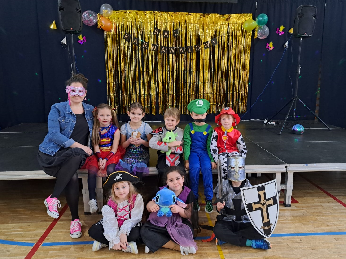 8 osobowa grupa dzieci w kolorowych kostiumach, siedzących na tle sceny ozdobionej złotą kurtyną z napisem „Bal Karnawałowy”. Każde dziecko jest przebrane za postać z bajki lub w fantazyjny strój, m.in. Superwoman, rycerza, pirata, Mario oraz strażaka. Obok dzieci siedzi uśmiechnięta kobieta w masce karnawałowej i jeansowej kurtce. W tle dekoracje wskazujące na okazję zabawy karnawałowej.
