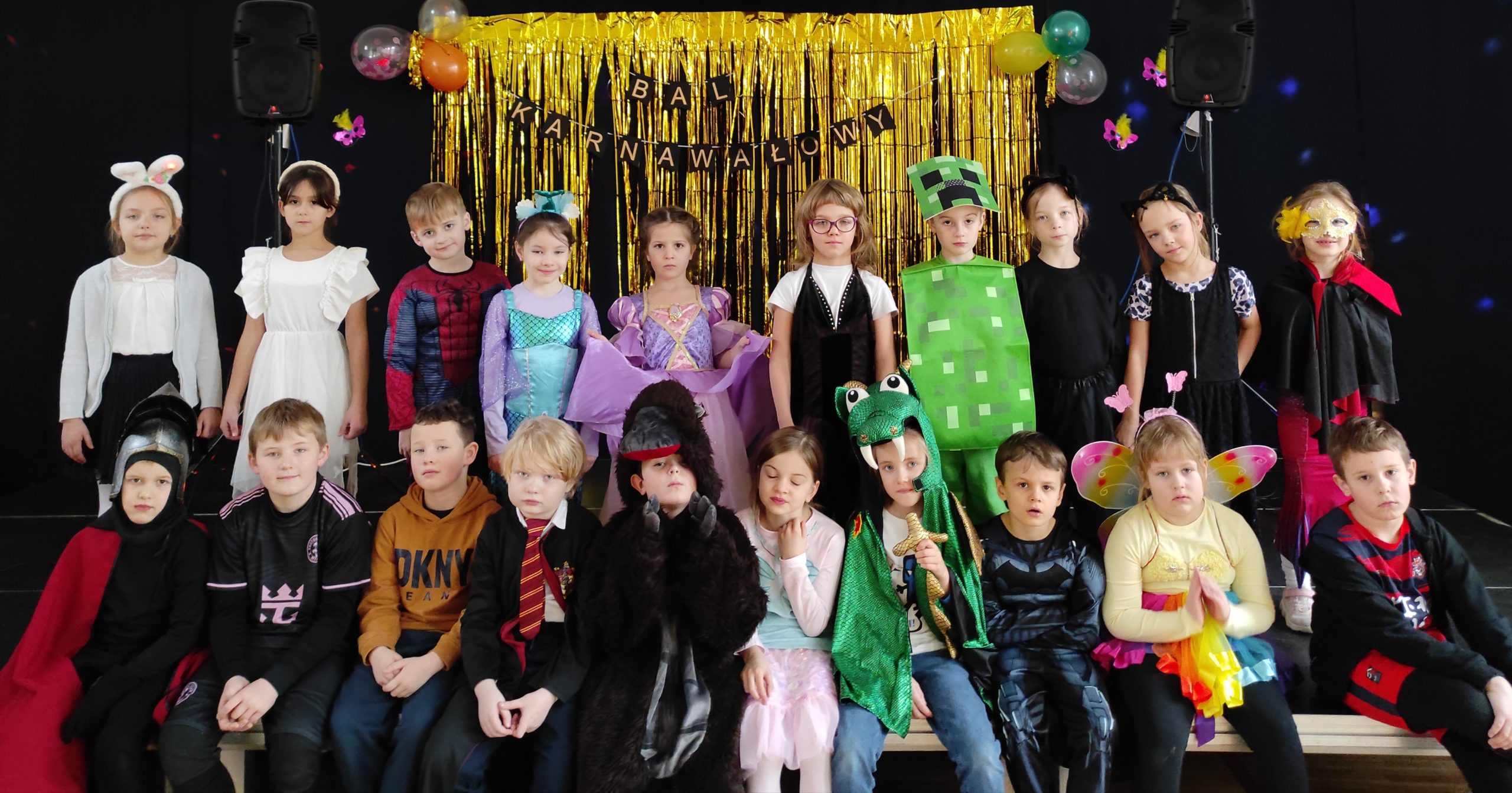 20 osobowa grupa dzieci, w strojach karnawałowych, ustawiona w dwóch rzędach, jeden stoi, drugi siedzi. W tle dekoracja karnawałowa, złota kurtyna i napis: BAL KARNAWAŁOWY.