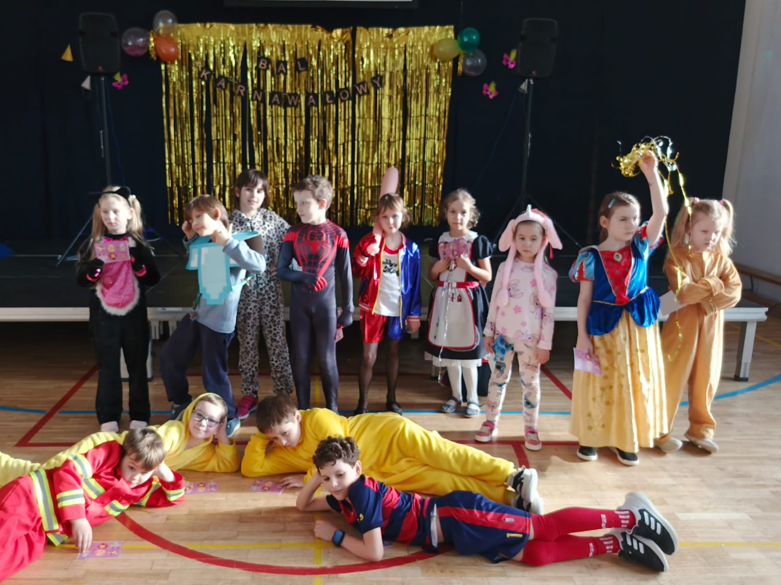 9 dzieci stoi w strojach karnawałowych, przed nimi leży 4 chłopców.