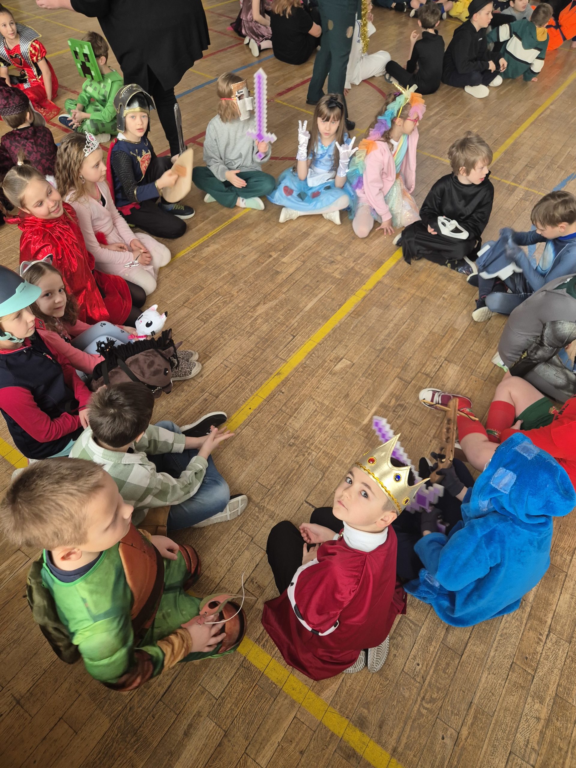 Dzieci siedzą na podłodze w kole. Jest ich 16. Dzieci są w strojach karnawałowych. Jeden chłopiec patrzy wysoko do góry. Ma na głowie koronę .