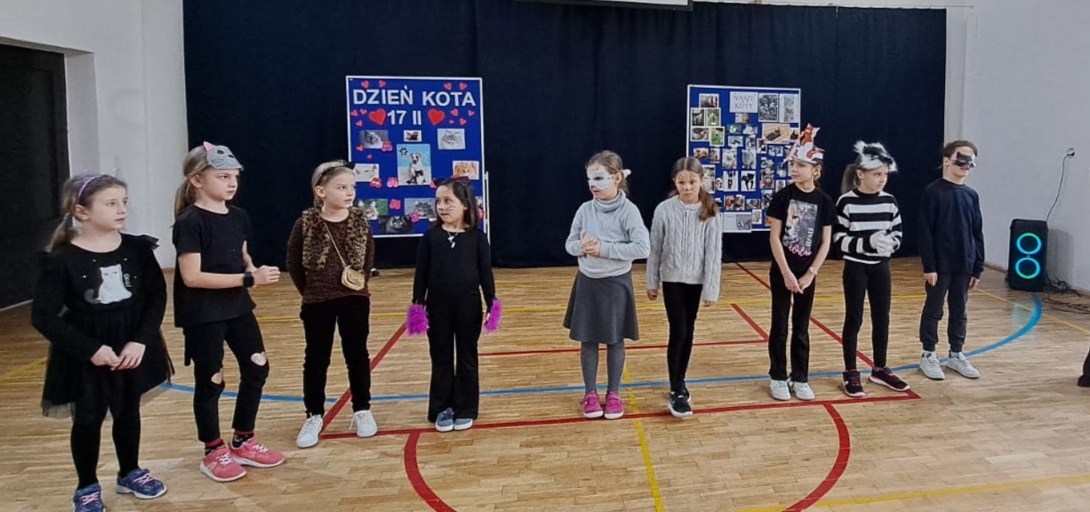 Na pierwszym planie 9 dzieci przebranych za koty, ustawionych w szeregu. W tyle granatowe tło oraz dwie tablice na niebieskim tle, jedna z napisem DZIEŃ KOTA, a druga z napisem NASZE KOTY.