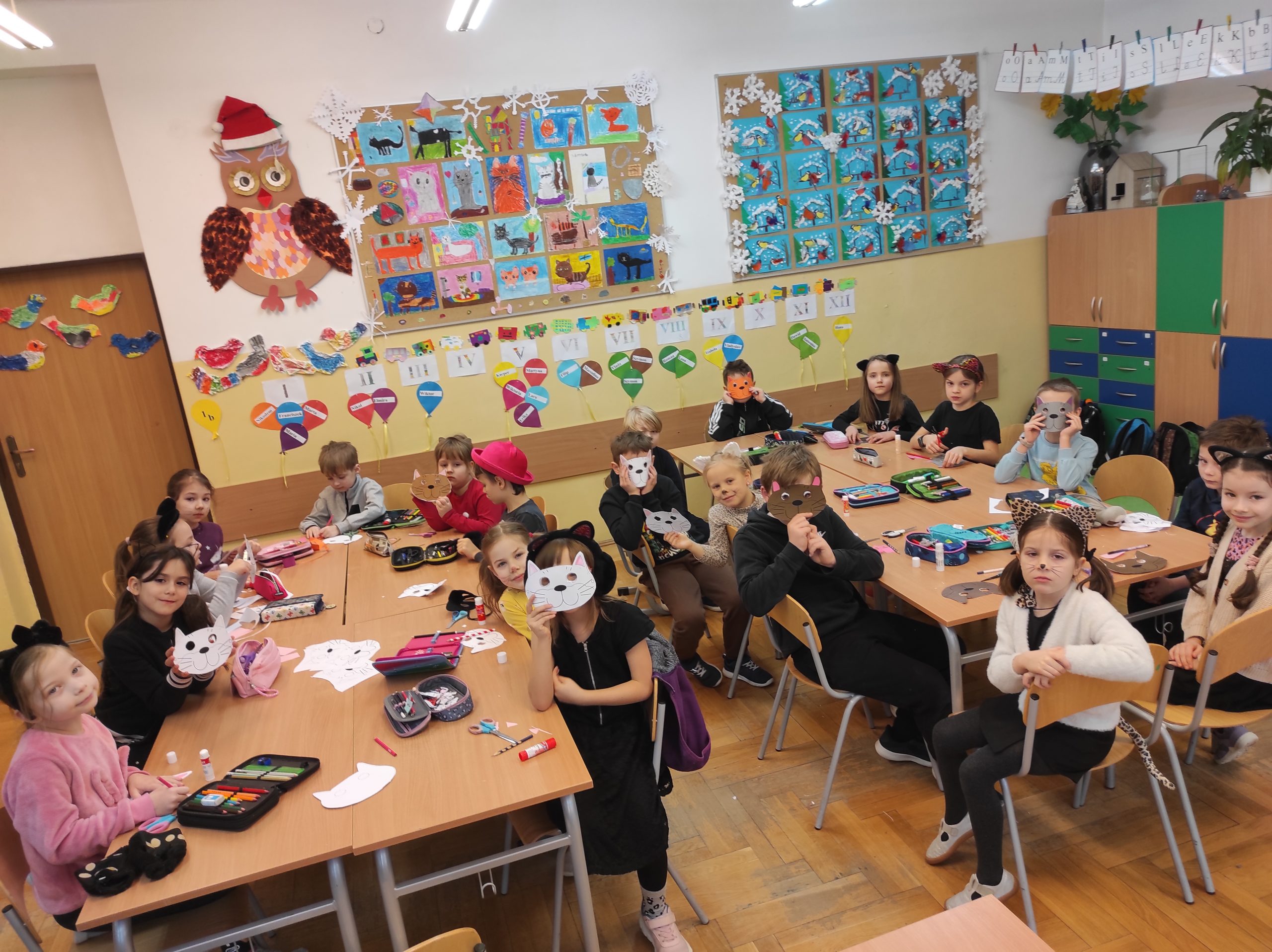 Sala lekcyjna, a w niej uczniowie siedzący na krzesłach, w dwóch grupach, przy zestawionych ze sobą stołach. Na ławkach piórniki, nożyczki, kleje, kolorowe maski kotów.