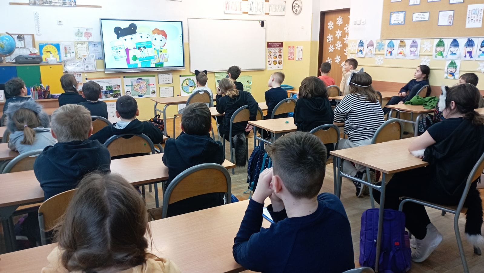 Sala lekcyjna, a w niej uczniowie siedzący w ławkach. Twarze dzieci skierowane są w stronę tablicy multimedialnej, na której wyświetlany jest obraz.