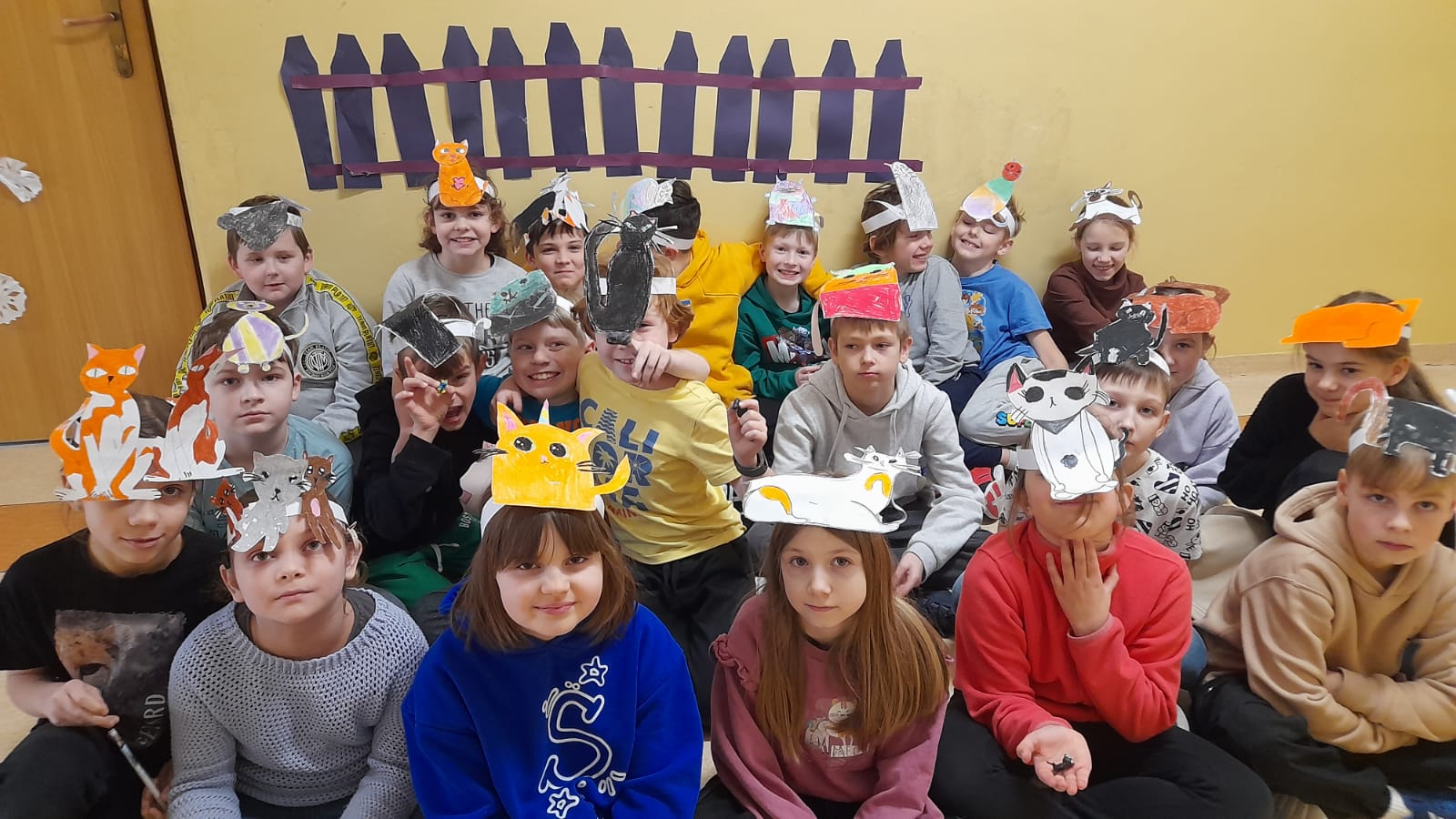 Uczniowie siedzą w trzech rzędach, na głowach mają opaski z kotami. W tle ściana, do której przyczepiony jest papierowy płotek.