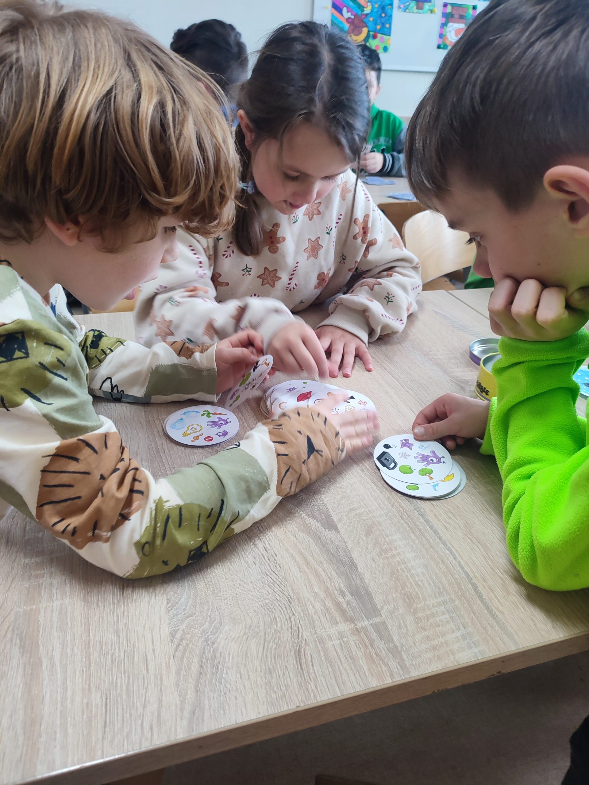 Trójka dzieci siedzi przy stoliku w ręku mają karty do gry w dobble.