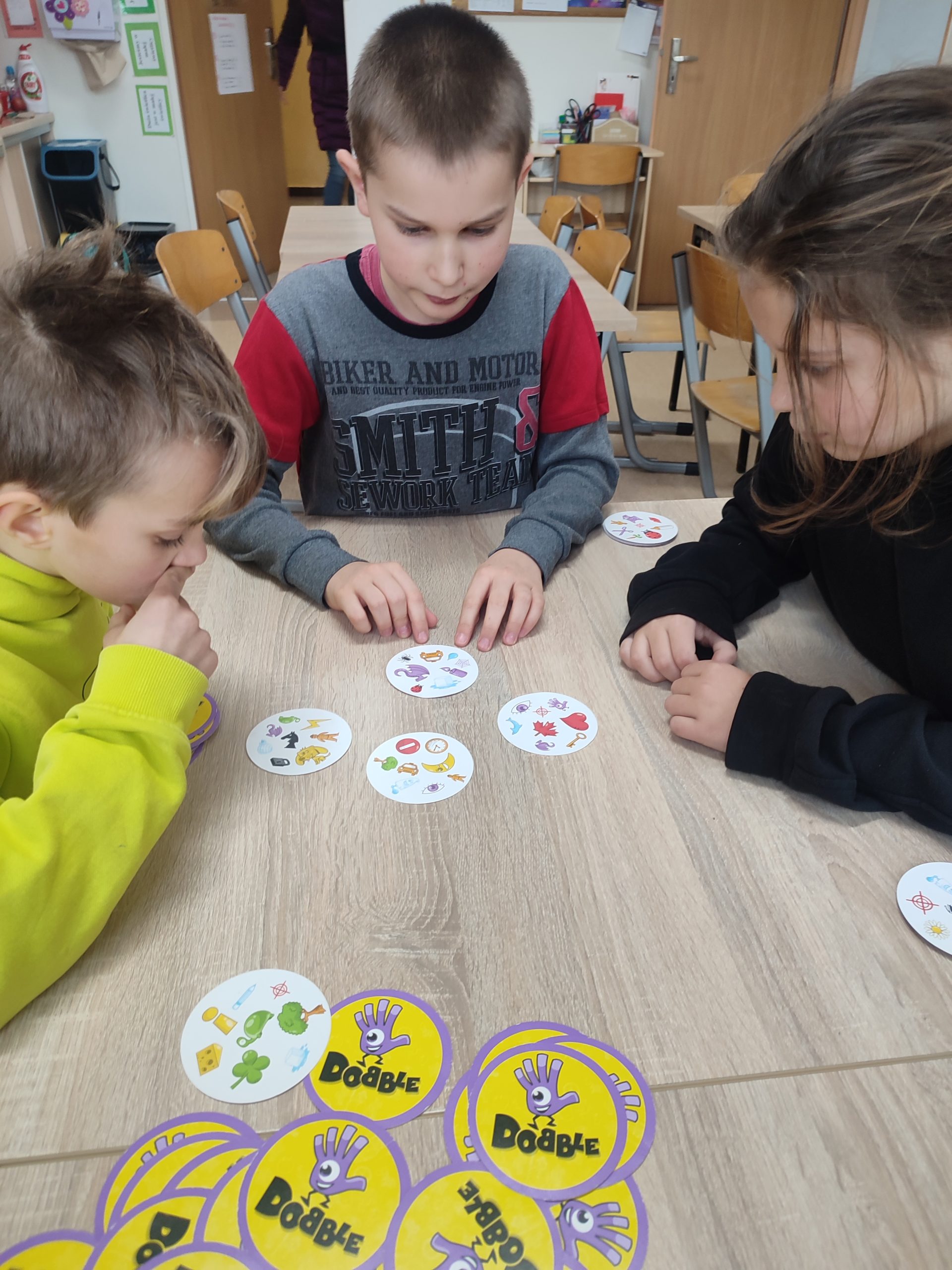 W świetlicy szkolnej trójka dzieci siedzi przy stoliku na którym leżą rozłożone karty do gry w dobble.