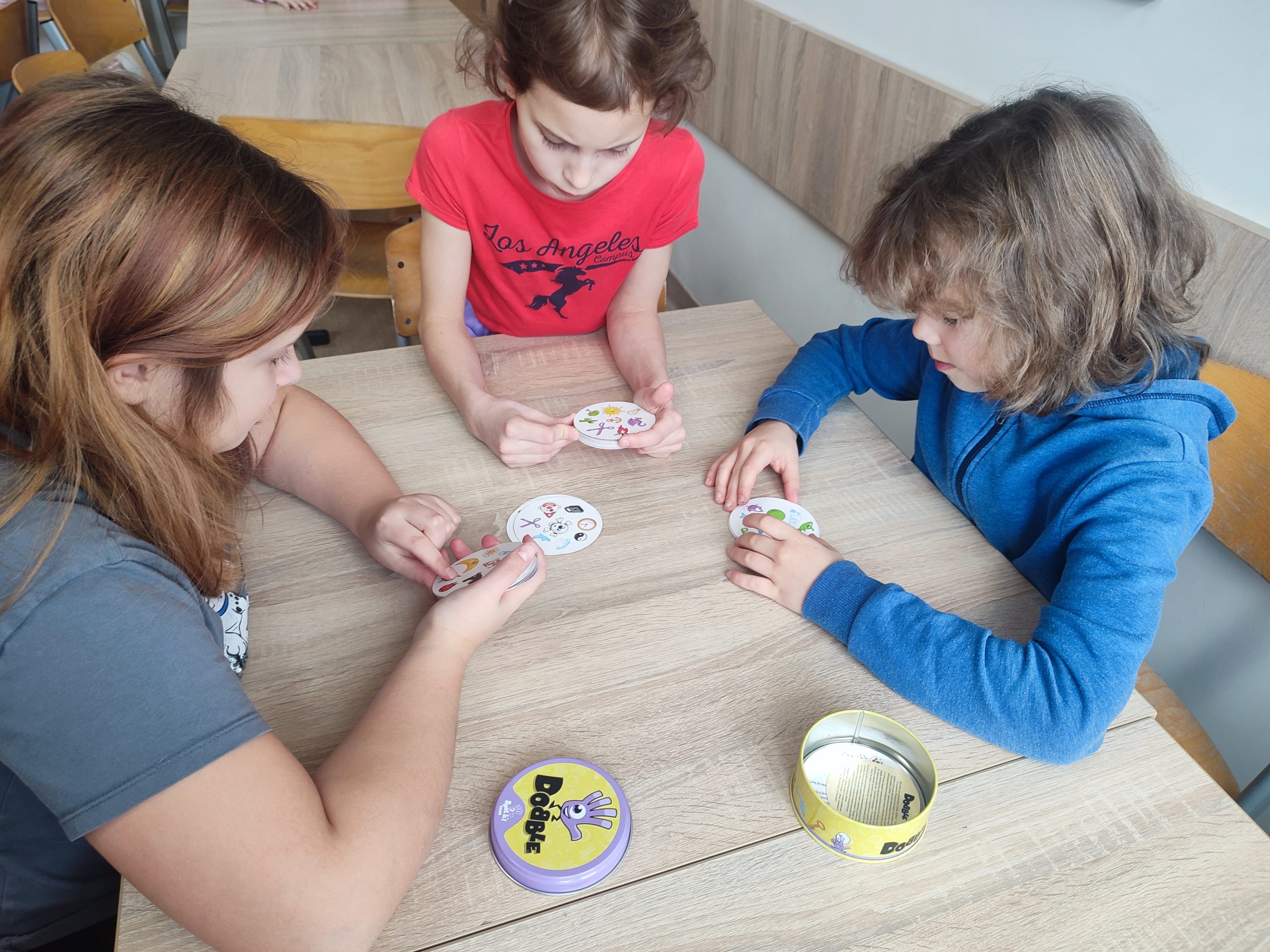 Trójka dzieci siedzi przy stoliku w ręku mają karty do gry w dobble.