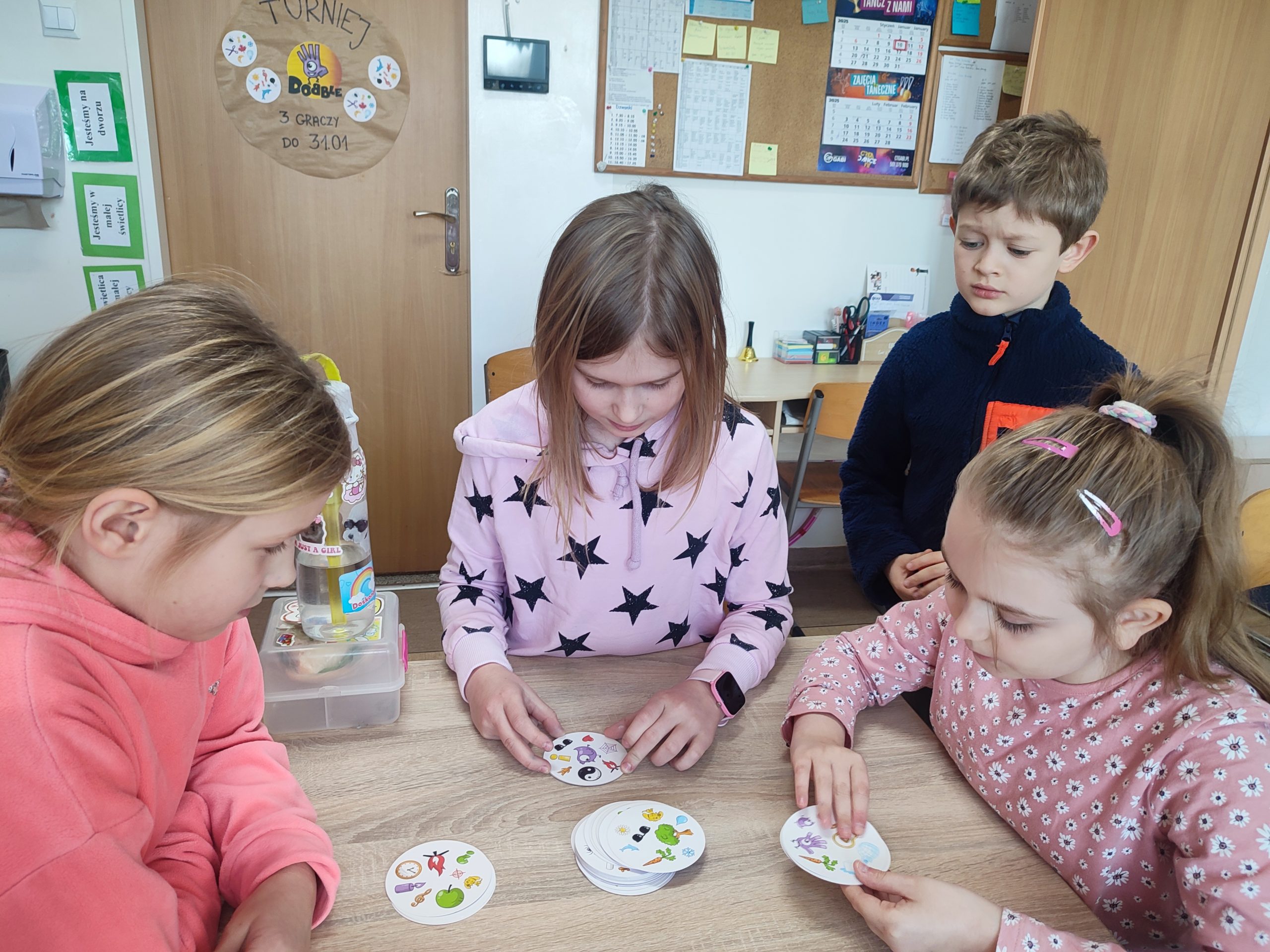 W świetlicy szkolnej trójka dzieci siedzi przy stoliku w  w ręku mają karty do gry w dobble. Obok stoi chłopiec i przygląda się grze.