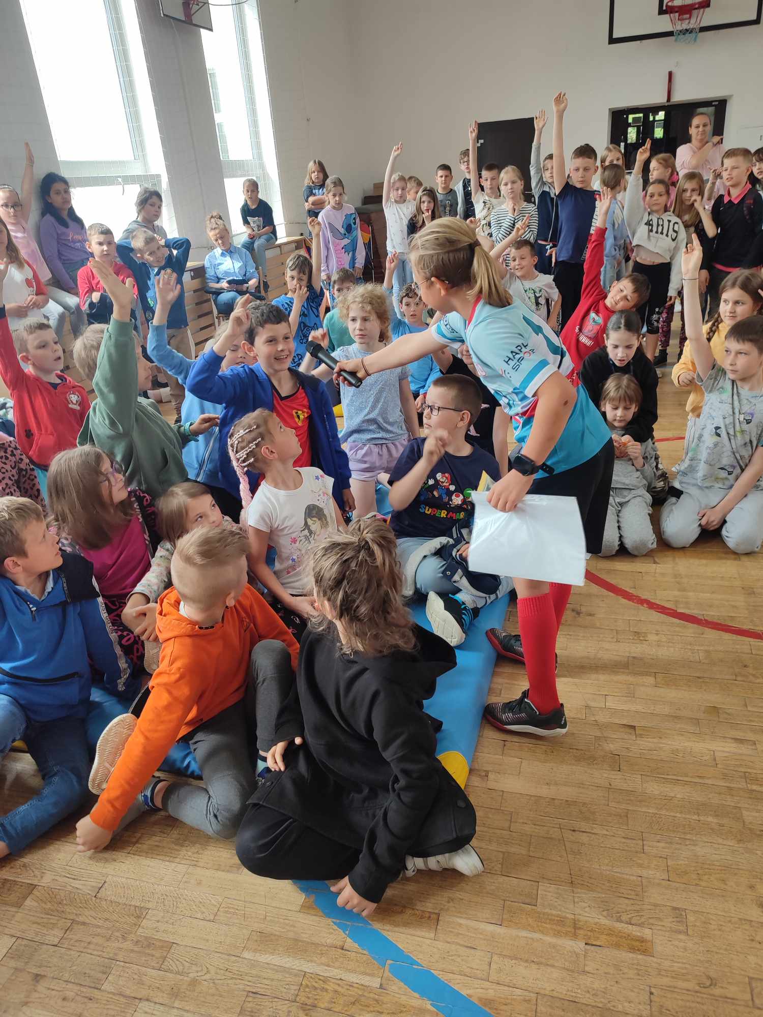 grupa dzieci zgromadzonych w sali gimnastycznej. Większość z nich siedzi na podłodze, a niektóre na ławkach przy ścianie. Wiele dzieci podnosi ręce do góry, chcąc odpowiedzieć na pytanie. W centrum kadru znajduje się dziewczyna w jasnoniebieskiej koszulce sportowej i czerwonych skarpetach, trzymająca mikrofon i kartkę, podchodząca do jednego z dzieci. W tle widać inne dzieci oraz nauczycieli obserwujących wydarzenie