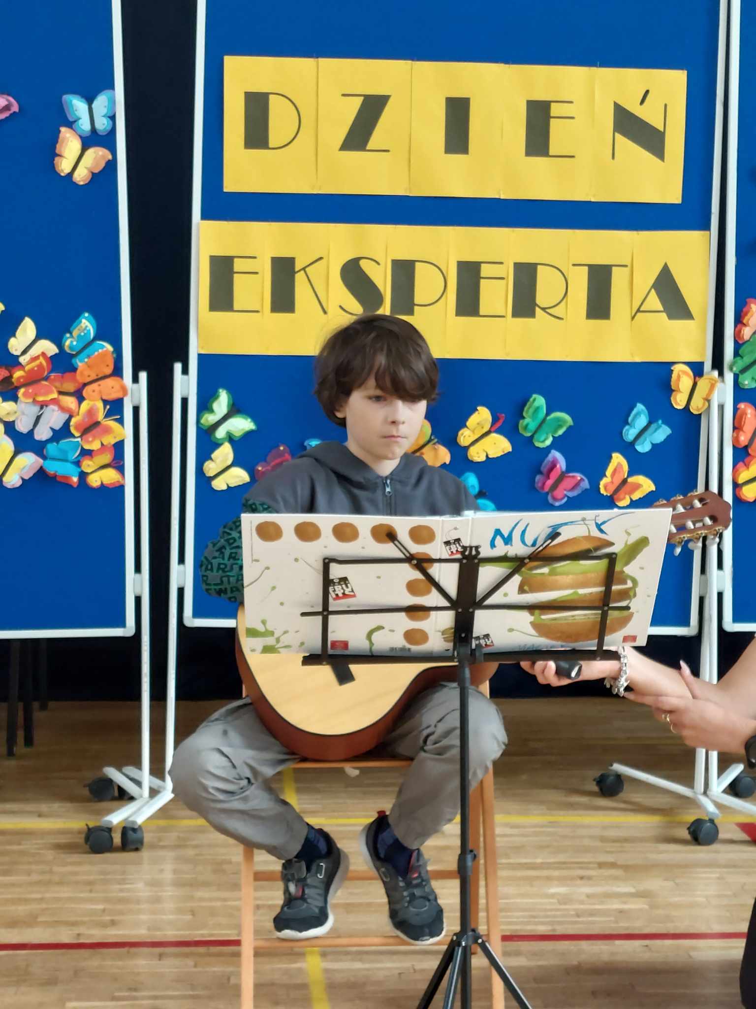 chłopiec z ciemnymi włosami, siedzący na wysokim stołku gra na gitarze. Przed nim stoi pulpit na nuty ozdobiony rysunkami, m.in. hamburgera. Chłopiec ma skupioną minę i patrzy na swoje nuty. W tle widać dekoracje z kolorowymi motylami oraz napis ‘DZIEŃ EKSPERTA’ na żółtym tle. Po prawej stronie zdjęcia dorosła osoba trzyma mikrofon w kierunku chłopca