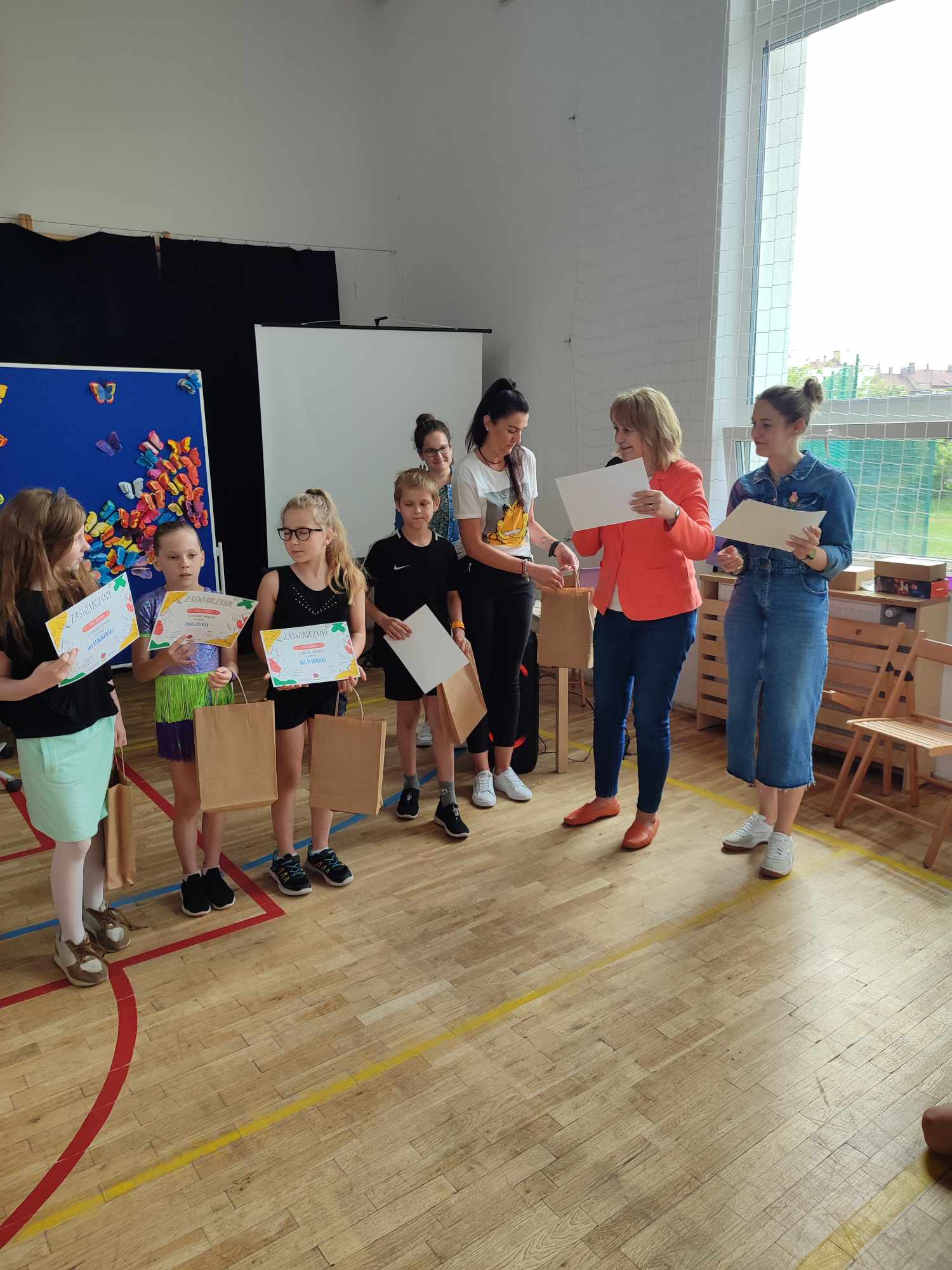 grupa dzieci i dorosłych w sali gimnastycznej podczas uroczystości wręczenia dyplomów i nagród. Dzieci stoją w rzędzie, trzymając kolorowe dyplomy oraz papierowe torby z upominkami. Po prawej stronie dwie kobiety wręczają im dokumenty i prowadzą ceremonię. W tle widać dekorację z kolorowymi motylami oraz ekran projekcyjny