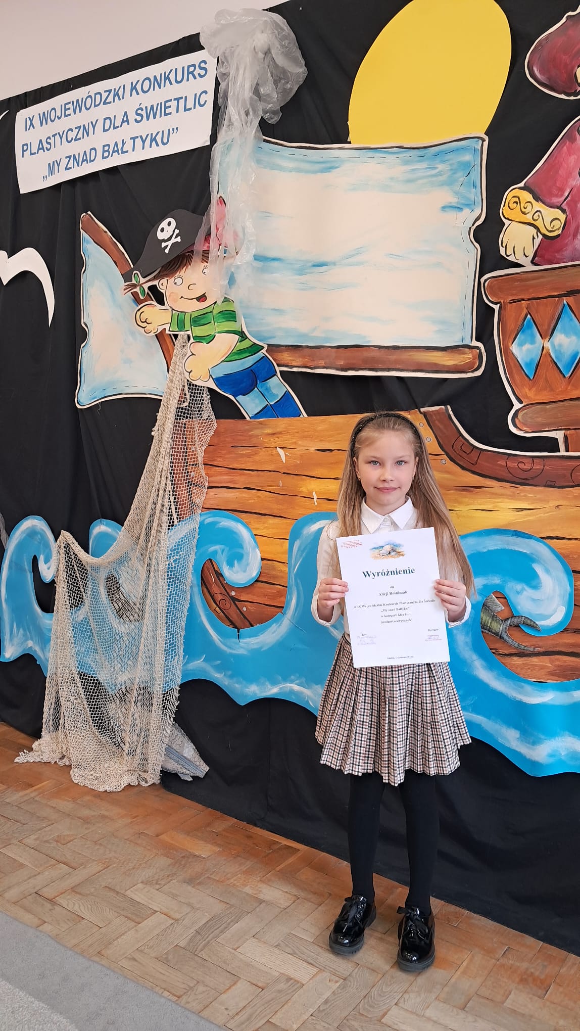 Dziewczynka trzymająca dyplom z wyróżnieniem stoi na tle dekoracji przedstawiającej piracki statek i fale morskie. W tle widnieje baner z napisem „IX Wojewódzki Konkurs Plastyczny dla Świetlic – My znad Bałtyku”.