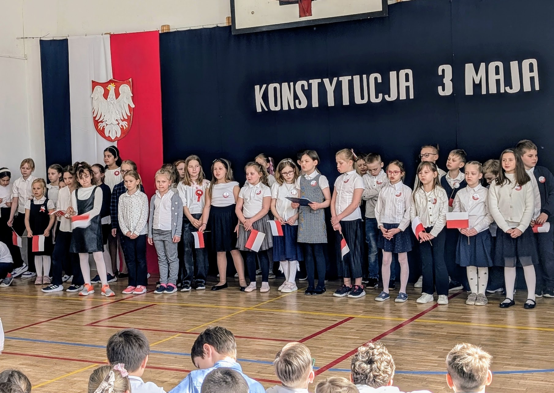 32 dzieci stoi obok siebie. Stoją w długim rzędzie. Dzieci ubrane są na galowo. Świętują święta majowe. Jedna dziewczynka trzyma w rękach teczkę i czyta. Za dziećmi jest granatowe tło, na którym znajduje się napis konstytucja 3 Maja. Po lewej stronie jest biało czerwona flaga na niej godło orzeł biały.