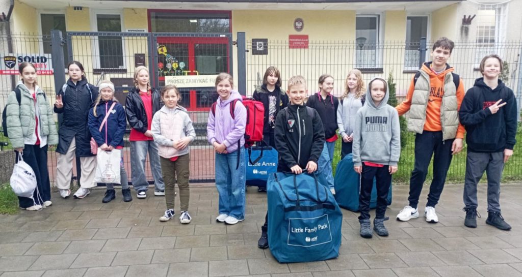 Na tle wejścia do budynku przedszkola stoi trzynastoosobowa grupa uczniów Koła HOPR. Wśród nich dziewczyna z apteczką i trójka uczniów dźwigających torby w fantomami.