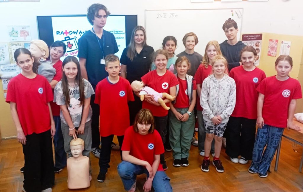 W sali lekcyjnej na tle tablicy stoi w dwóch rzędach grupa uczniów Koła HOPR wraz z dwoma studentami medycyny GUMed. Troje uczniów trzyma fantomy.