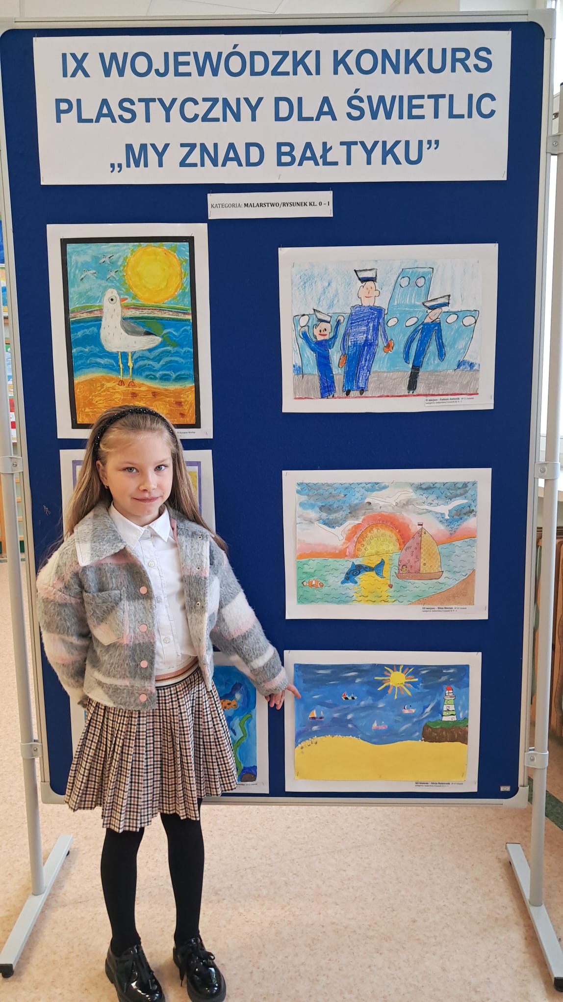 Dziewczynka wskazuje palcem na swoją pracę plastyczną przedstawiającą nadmorski krajobraz z latarnią morską i żaglówkami. W tle plansza z pracami konkursowymi oraz baner z napisem „IX Wojewódzki Konkurs Plastyczny dla Świetlic – My znad Bałtyku”.