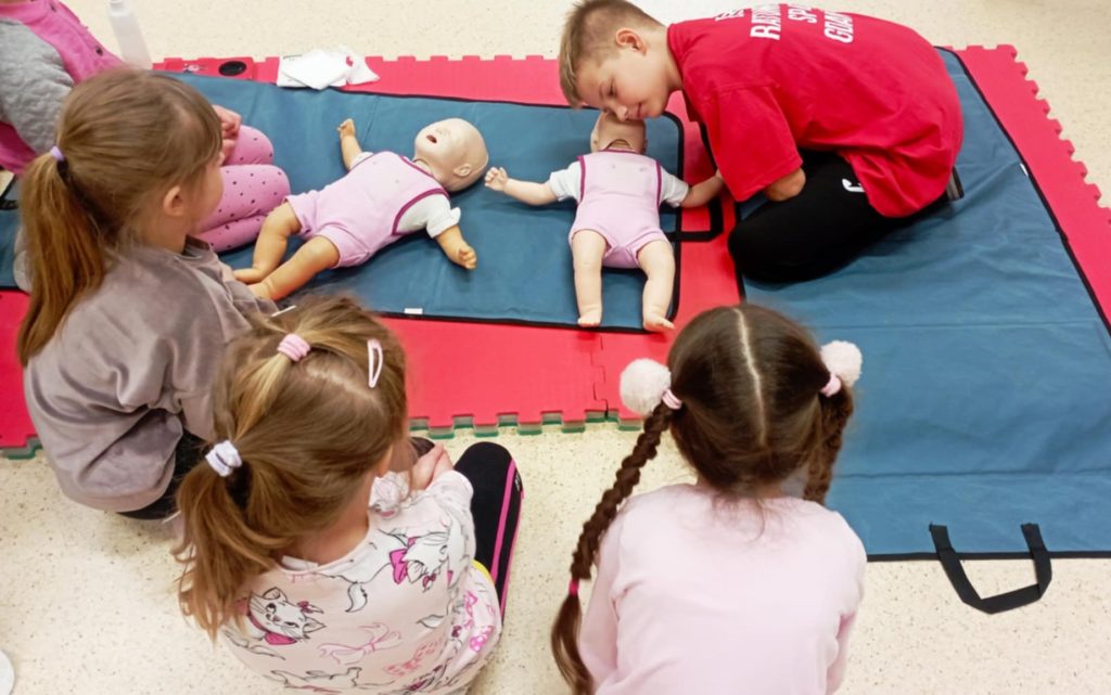 Na pierwszym planie uczeń Koła HOPR nachylający się nad fantomem niemowlęcia, obok leży drugi fantom. Przed uczniem siedzą cztery przedszkolaki.