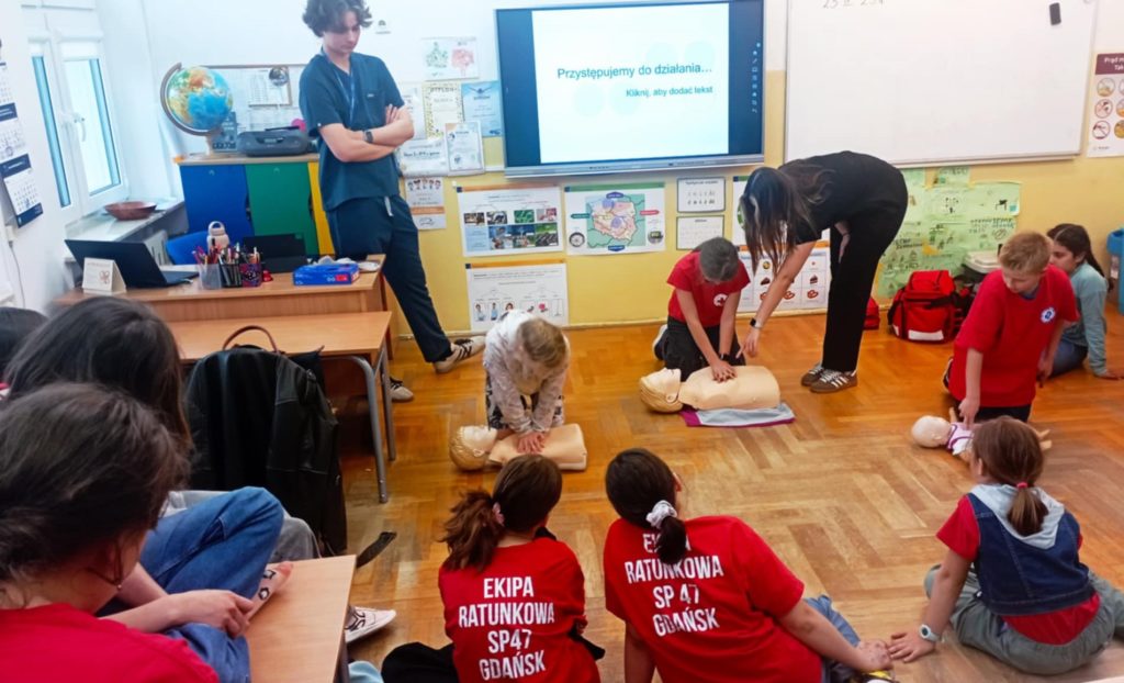 W sali lekcyjnej na tle tablicy multimedialnej z napisem „Przystępujemy do działania” na pierwszym planie 3 uczniów Koła HOPR uciskających klatkę piersiową fantomów. Przy nich stojący studenci medycyny, w koło pozostali uczniowie.