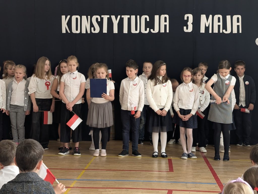 15 dzieci stoi obok siebie. Jedna dziewczynka jest z przodu wszystkich. Czyta tekst apelowy. Dziewczynka ta ma na sobie rajstopy spódniczkę i białą bluzkę. Nad dziećmi jest napis Konstytucja 3 Maj. Napis ten wisi na granatowych płótnach.