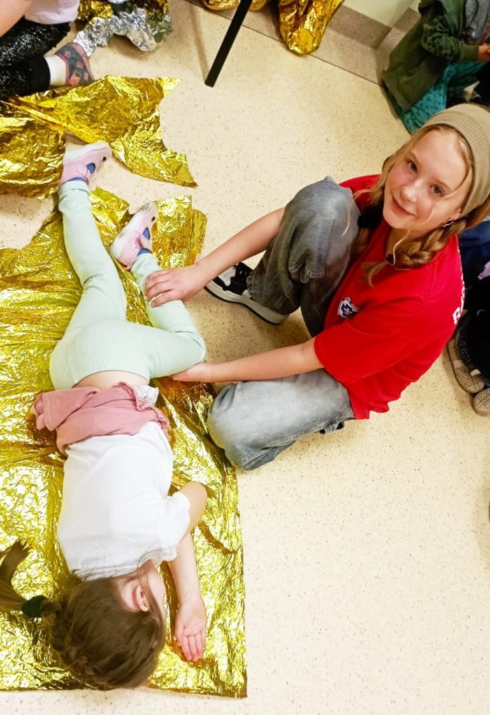 Na folii NRC leży dziewczynka ułożona w bocznej pozycji. Obok niej klęczy uczennica Koła HOPR.