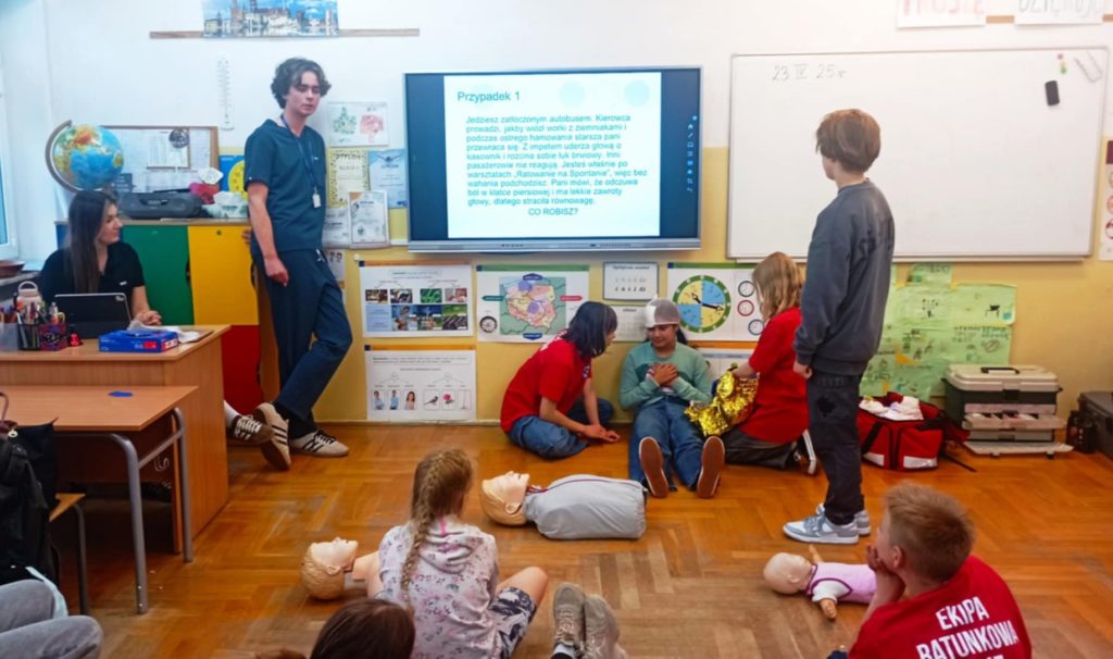 W sali lekcyjnej na tle tablicy multimedialnej z napisem „Przypadek 1” na pierwszym planie 4 uczniów Koła HOPR biorących udział w pozoracji wypadku. Przy tablicy 2 studentów, w koło inni uczniowie.