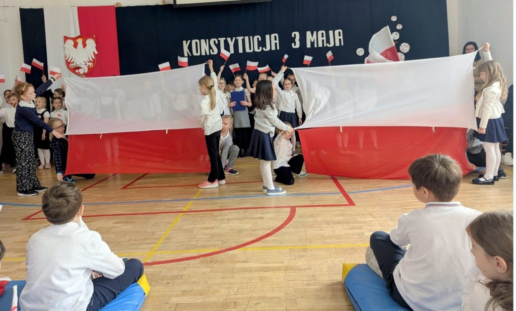 Dwoje dzieci trzyma flagę po prawej stronie patrząc, po lewej stronie patrząc również dwoje dzieci trzyma flagę. Dzieci wykonują taniec z flagą Polski. Dzieci są ubrane na galowo. Za dziećmi stoją kolejne dzieci, które w rękach mają chorągiewki z barwami flagi polskiej. Dzieci nimi