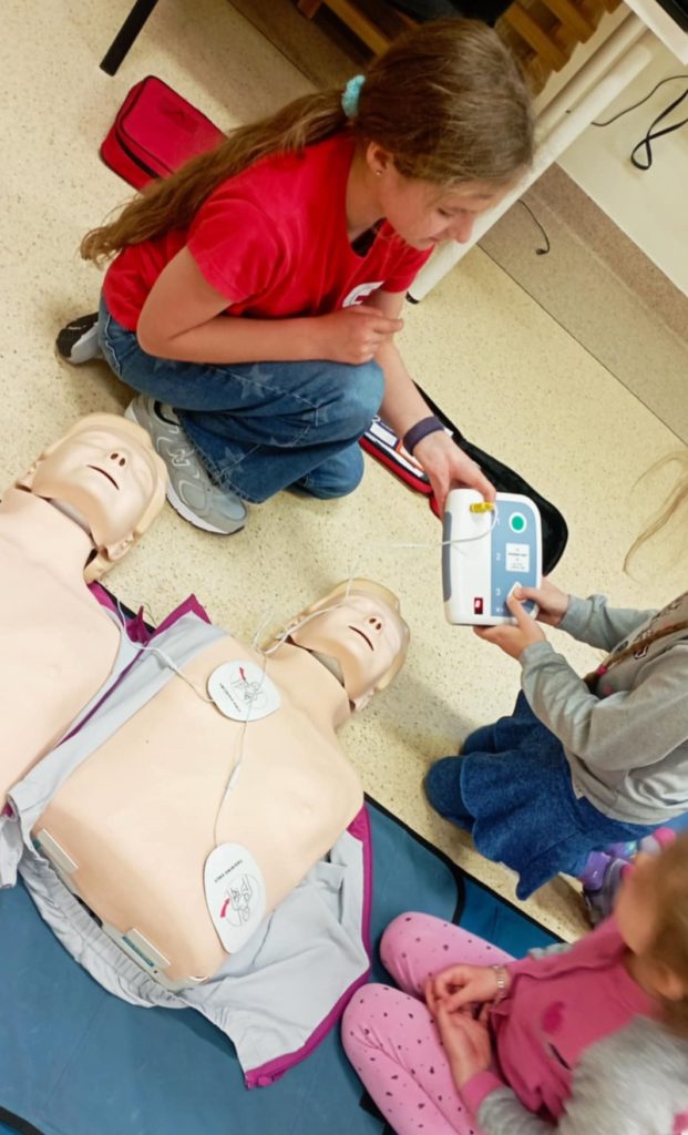 Na pierwszym planie leżą dwa fantomy, jeden ma podłączone elektrody AED. Za nimi uczennica Koła HOPR trzymająca w dłoni defibrylator treningowy. Obok dwie dziewczynki, jedna z nich wciska przycisk AED.