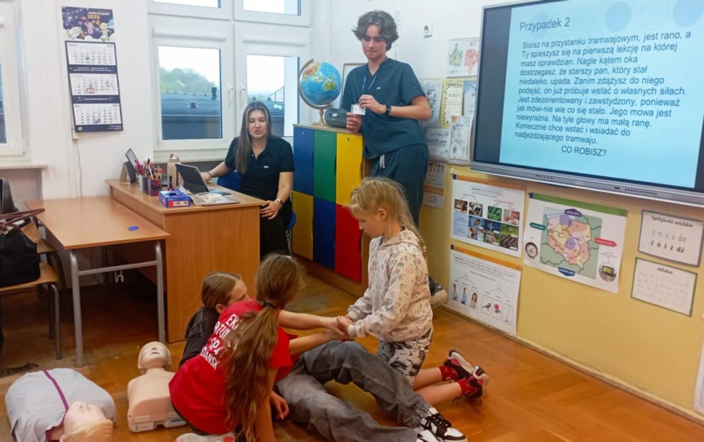 W sali lekcyjnej na tle tablicy multimedialnej z napisem „Przypadek 2” na pierwszym planie 3 uczniów Koła HOPR biorących udział w pozoracji wypadku. Przy tablicy 2 studentów, w koło leżące fantomy.