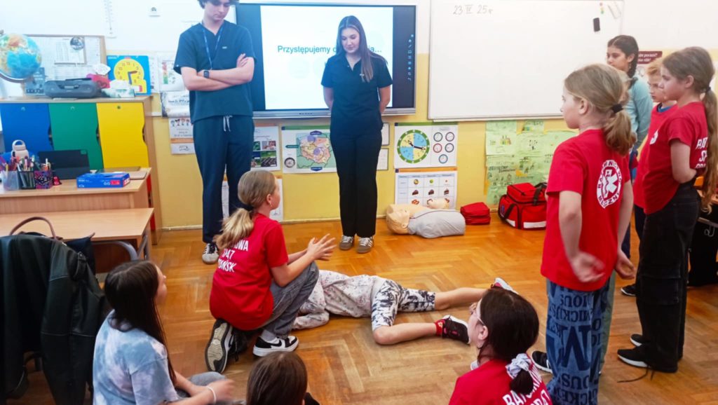 W sali lekcyjnej na tle tablicy multimedialnej z napisem „Przystępujemy do działania” na pierwszym planie 2 uczniów Koła HOPR biorących udział w pozoracji wypadku. Przy tablicy 2 studentów, w koło inni uczniowie.