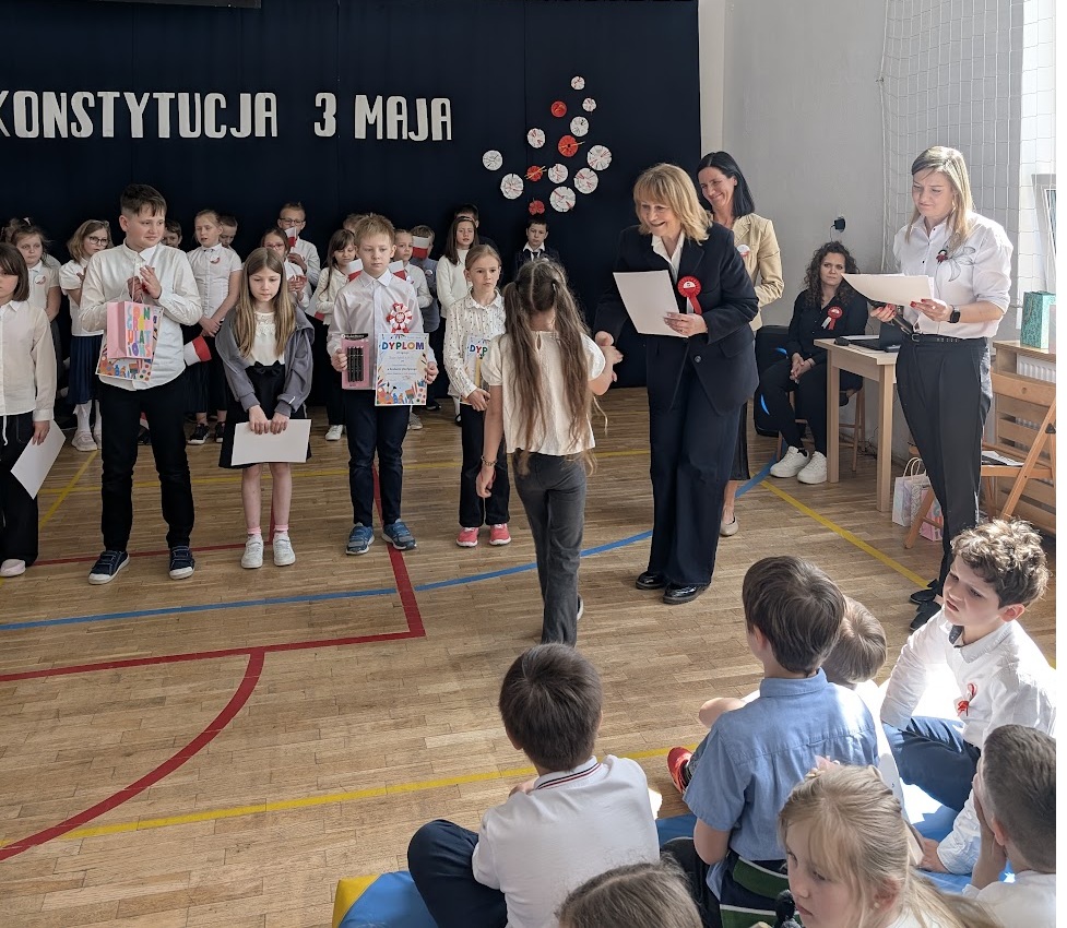 Grupa dzieci stoi z dyplomami i nagrodami w rękach. Dzieci stoją w rzędzie obok dzieci stoi Pani, która daje dziecku - dziewczynce dyplom. Pani dyrektor gratuluje dziewczynce. Za Panią która wręcza dyplom po prawej i po lewej stronie stoją dwie Panie a za tymi Paniami jedzie przy stoliku jeszcze jedna Pani.