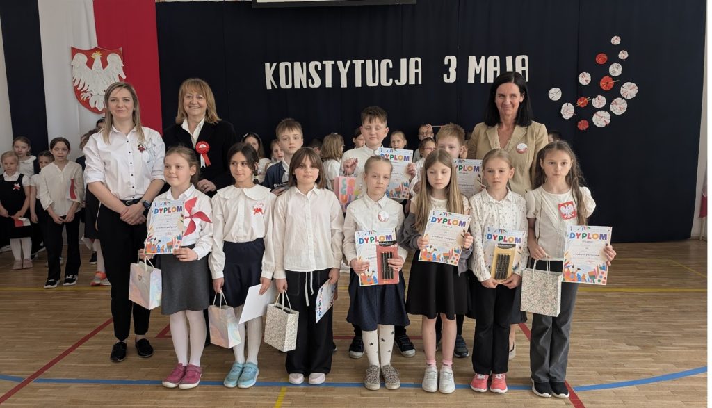 Grupa dzieci stoi obok siebie. Dzieci jest 10. Dzieci są ubrane na galowo. Dzieci trzymają w rękach dyplomy z okazji udziału w konkursie plastycznym. Po Lewej stronie dzieci stoi=ją dwie Panie .Po prawej stoi jedna Pani. Za dziećmi i Paniami jest płótno na tle którego widnieje napis Konstytucja 3 Maj.
