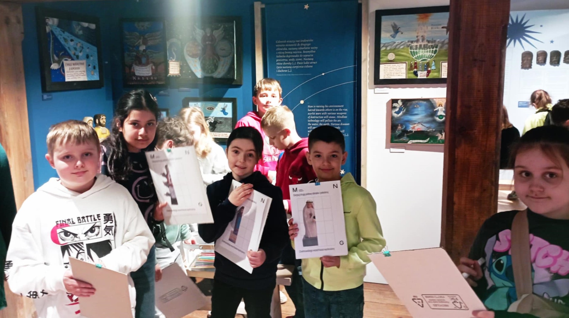 Na tle wyeksponowanych obrazów wystawy „Chełmowskiego wszechświat wyobrażony” stoją uczniowie klasy 3c trzymający w dłoniach karty pracy z zadaniami lekcji muzealnej