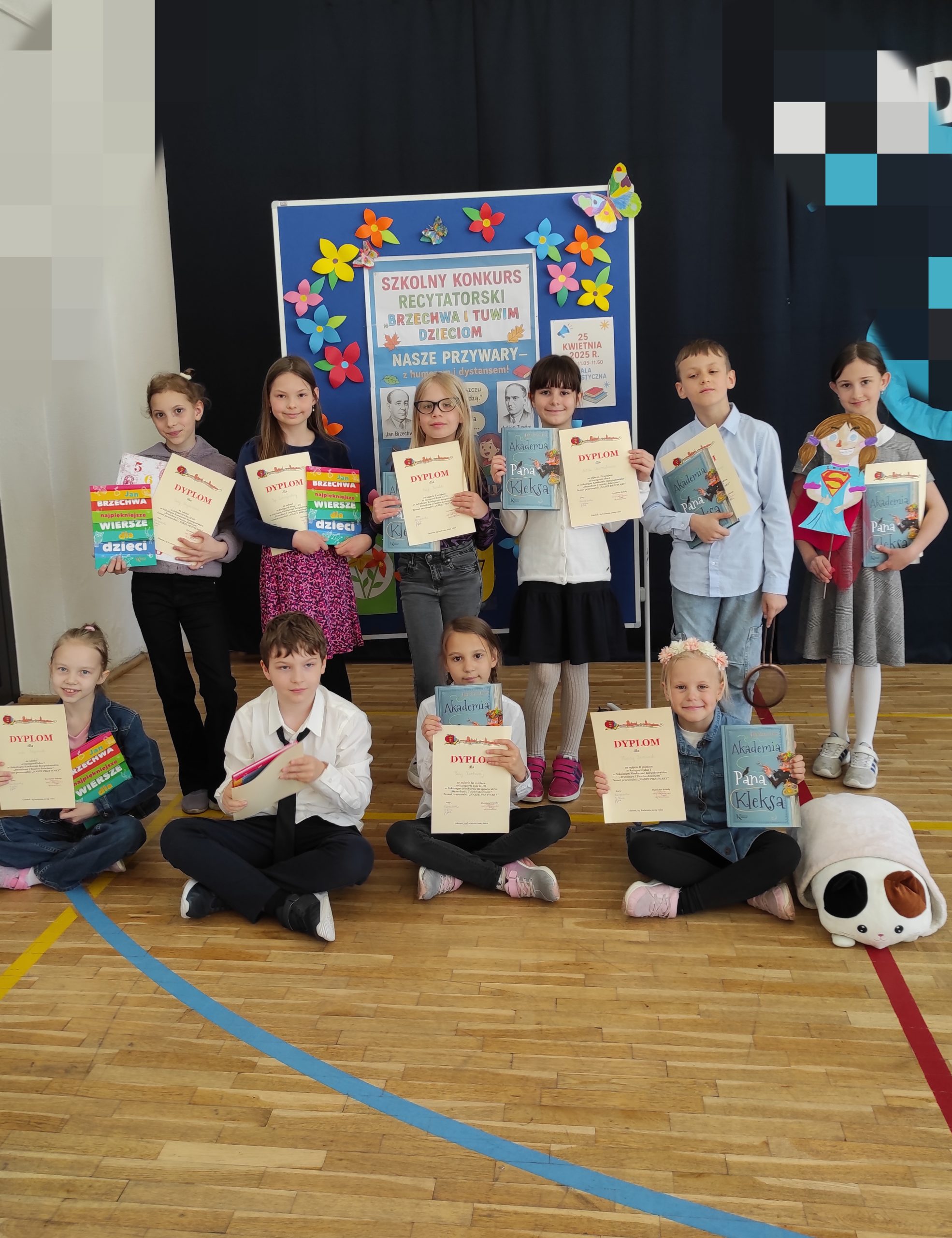 10 uczniów ustawionych w 2 rzędach, jeden siedzi, drugi stoi. Dzieci trzymają w dłoniach dyplomy i książki. W tle tablica informacyjna z napisem: KONKURS RECYTATORSKI "Brzechwa i Tuwim dzieciom" - NASZE PRZYWARY i kolorowymi kwiatami.