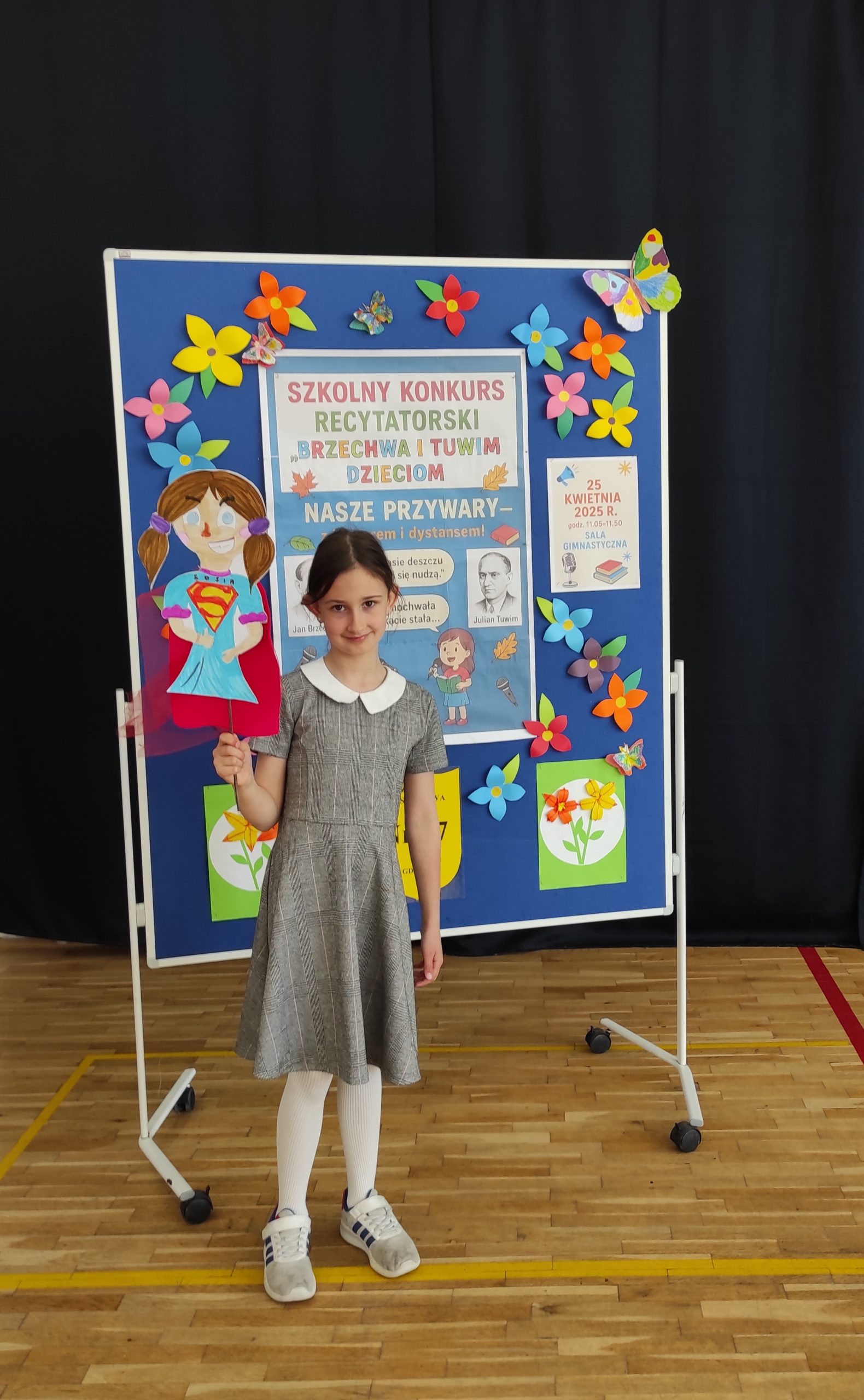 W tle tablica informacyjna z napisem: KONKURS RECYTATORSKI "Brzechwa i Tuwim dzieciom" - NASZE PRZYWARY i kolorowymi kwiatami. Na pierwszym planie stoi dziewczynka, która trzyma w dłoni kukiełkę przedstawiającą dziewczynkę.