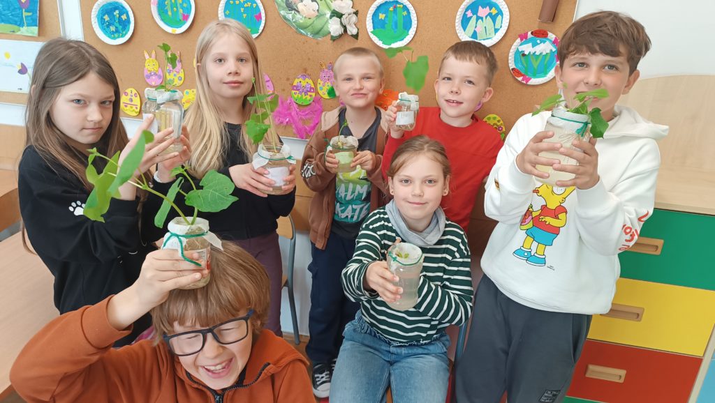 Grupa uśmiechniętych dzieci w klasie trzyma sadzonki roślin w słoikach z wodą.  Dzieci są podekscytowane i dumne ze swoich roślin. W tle widoczna jest kolorowa tablica z pracami plastycznymi.