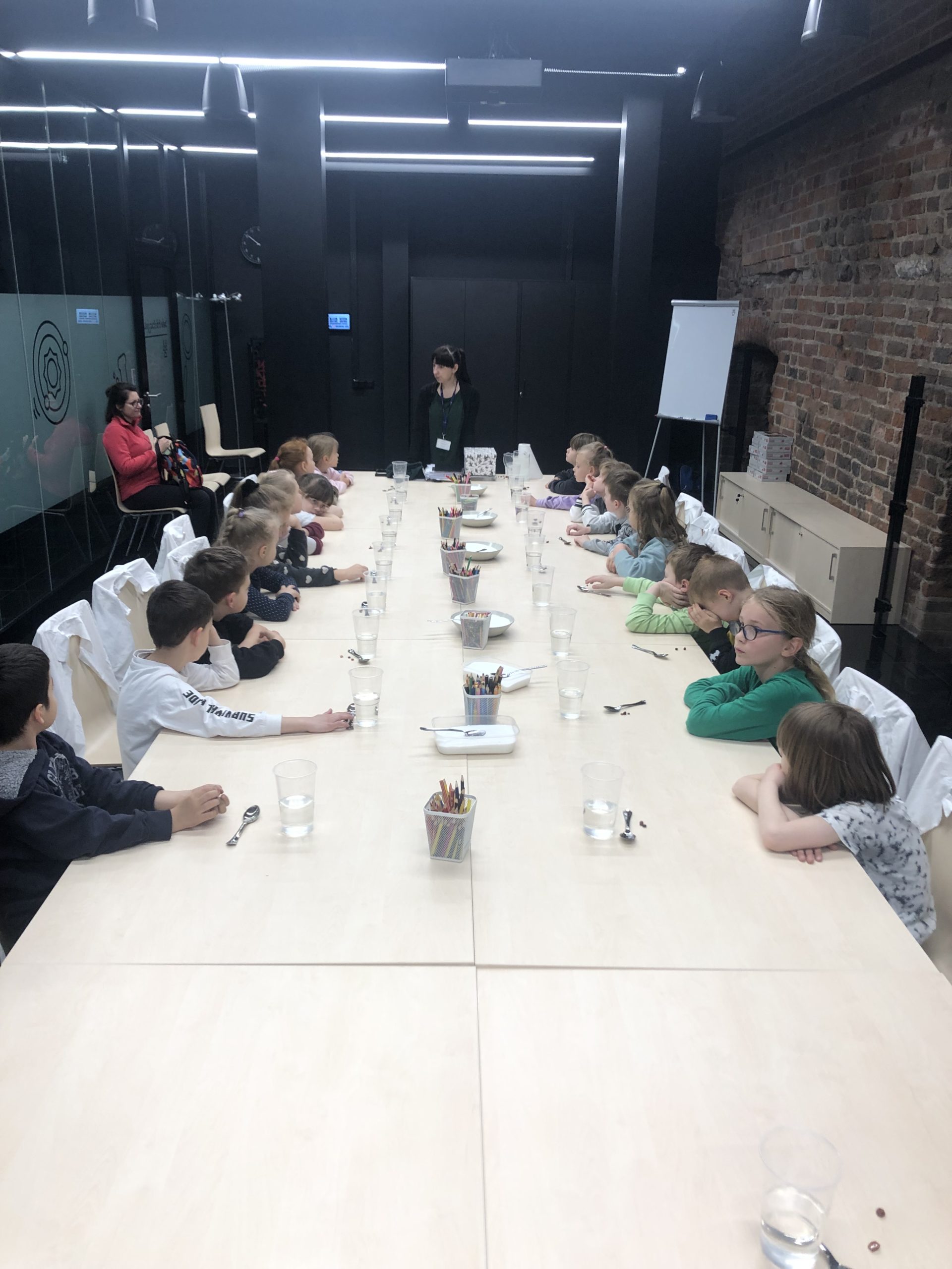 18 dzieci siedzi przy stole, na końcu stołu stoi pani. Na stole leżą kredki, kubki z wodą, łyżeczki bursztyny.