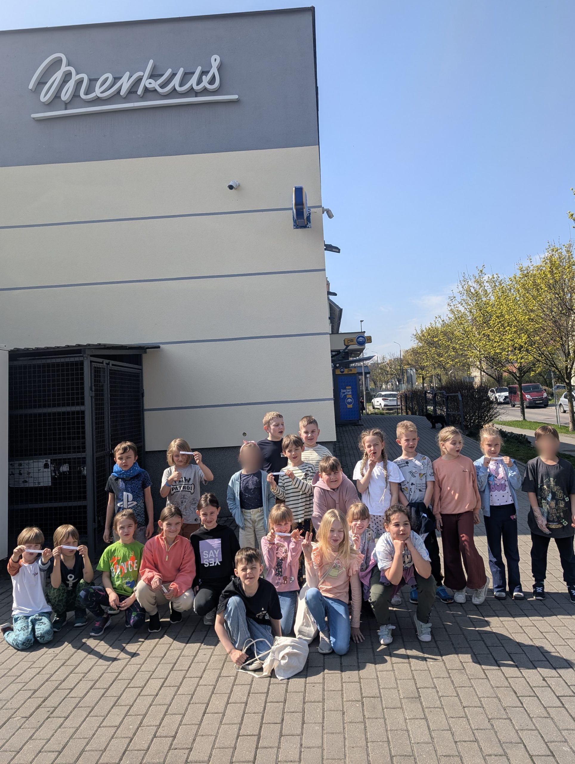 Grupa uśmiechniętych dzieci z klasy 1 pozuje do zdjęcia na tle budynku sklepu Merkus. Pogoda jest słoneczna, a dzieci stoją lub kucają w dwóch rzędach, gotowe na wspólne zakupy.
